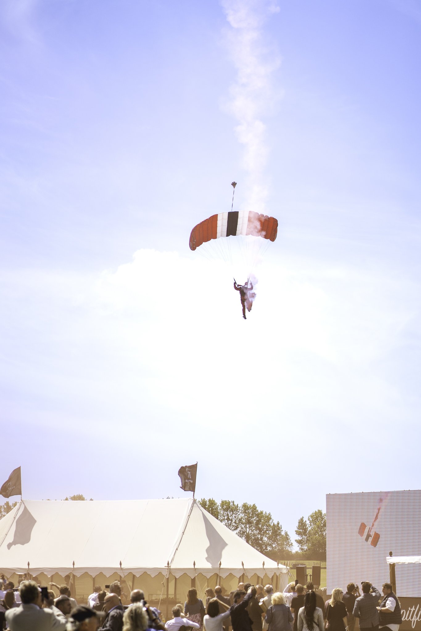 Parachutist descending with red, white, blue, and black parachute over a crowd gathered outside a large white tent at an outdoor event.