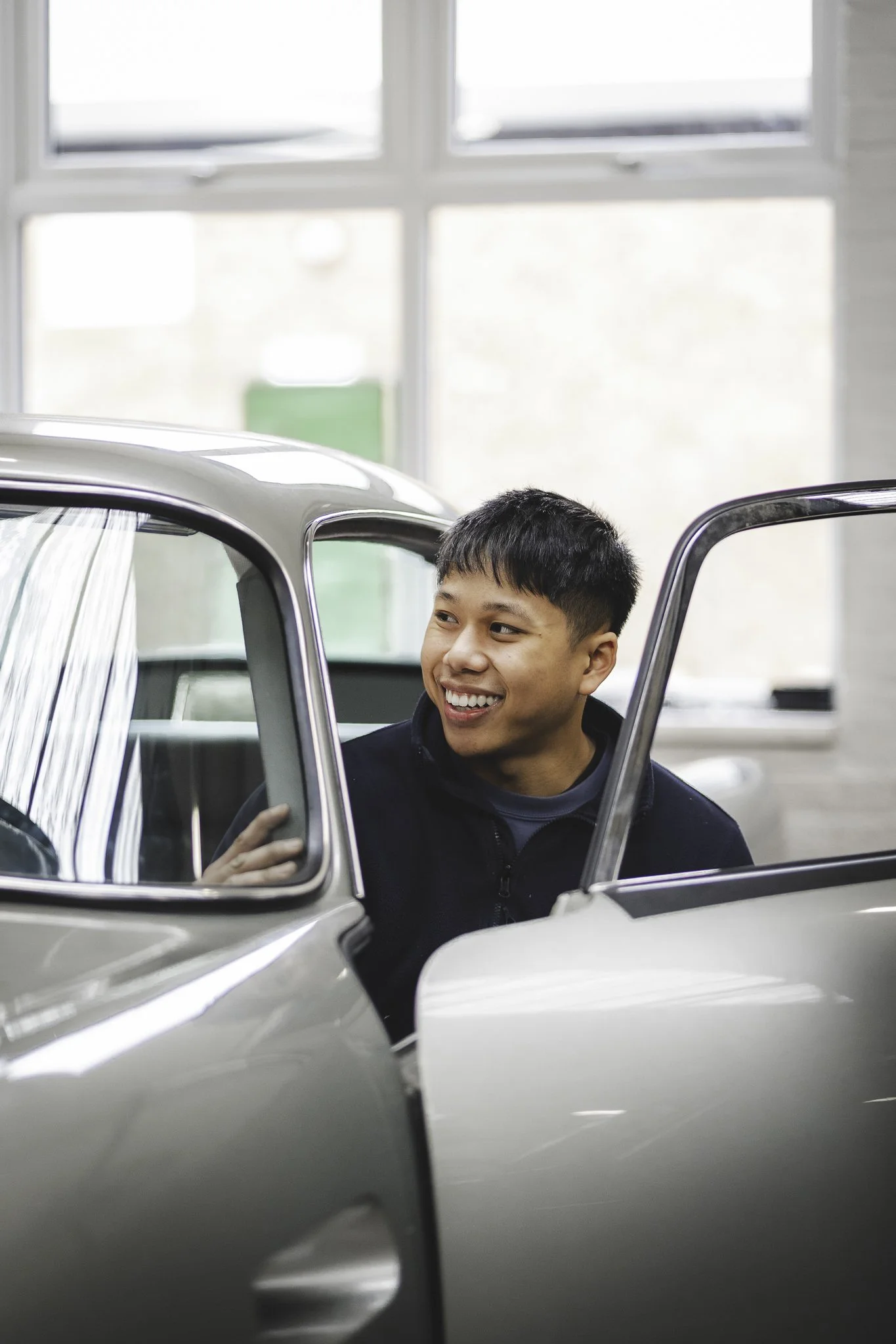 A young man smiling while sitting inside a vintage silver car with the door open, in a bright indoor space with large windows.