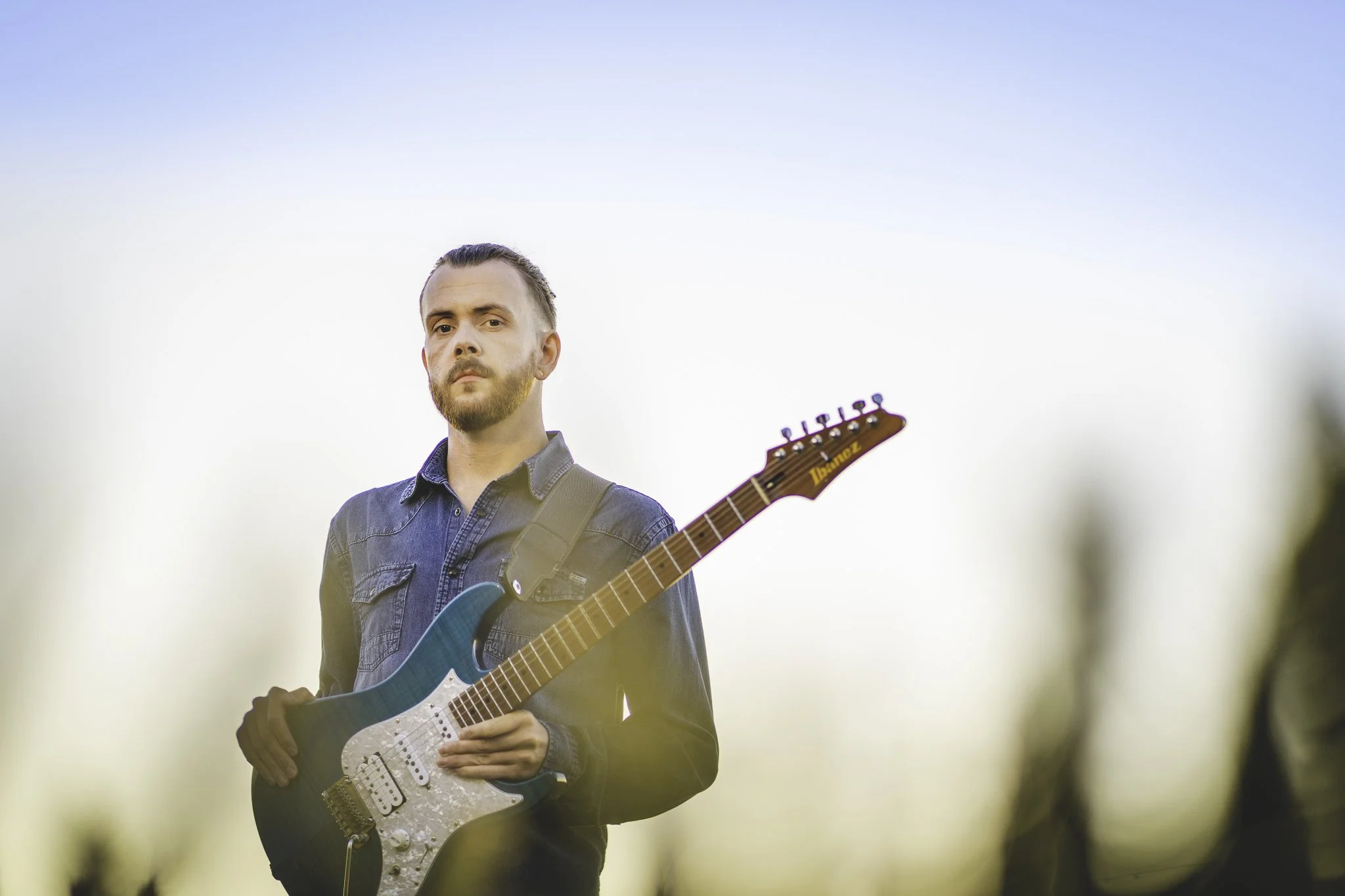 A man with a beard and mustache standing outdoors at sunset, holding an electric guitar, dressed in a denim shirt, with blurred trees in the background.