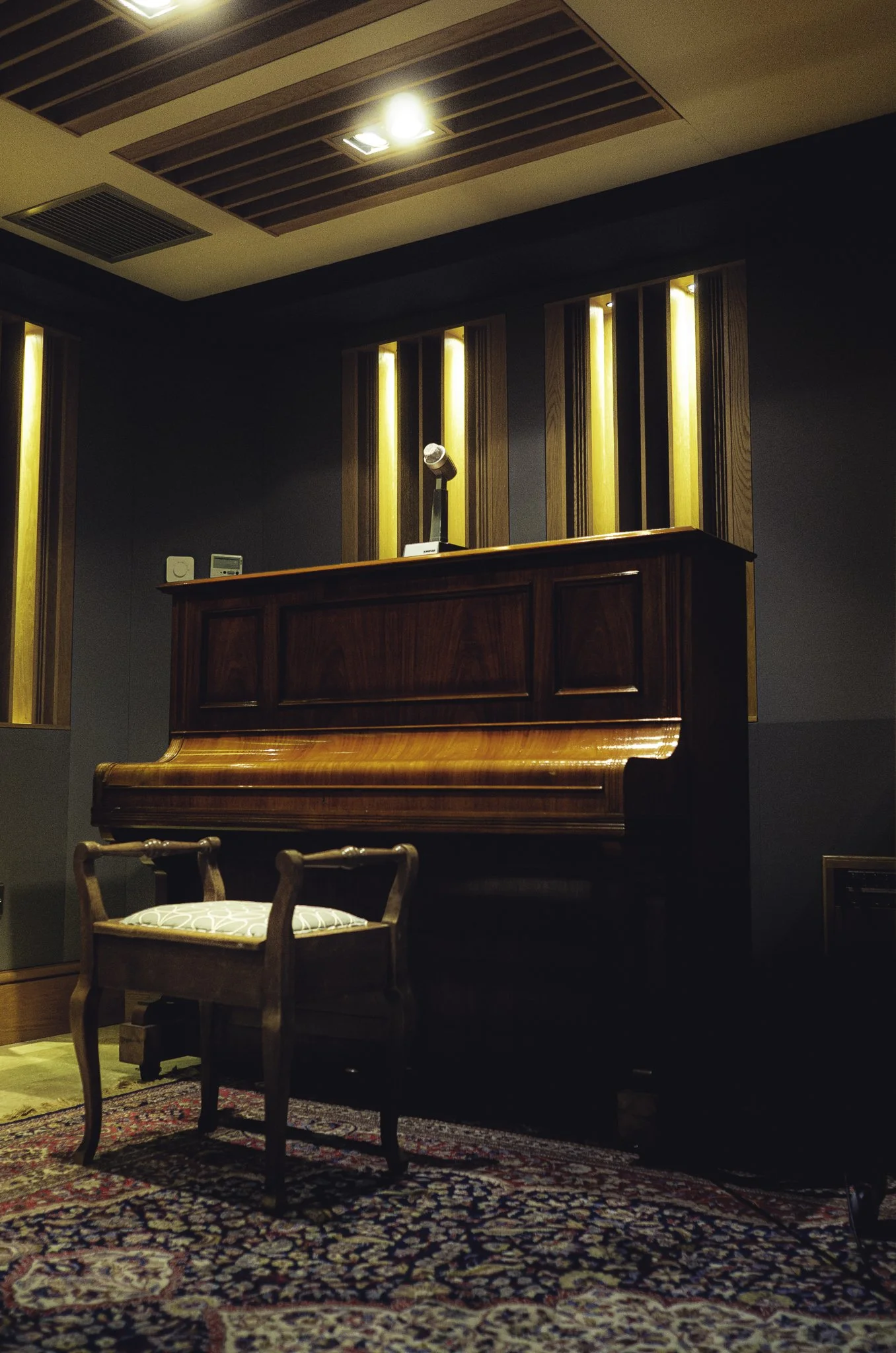 A recording studio room with a polished wooden upright piano, a vintage wooden chair with cushioned seat in front of it, decorative wooden accent panels with warm lighting, and a patterned rug on the floor.