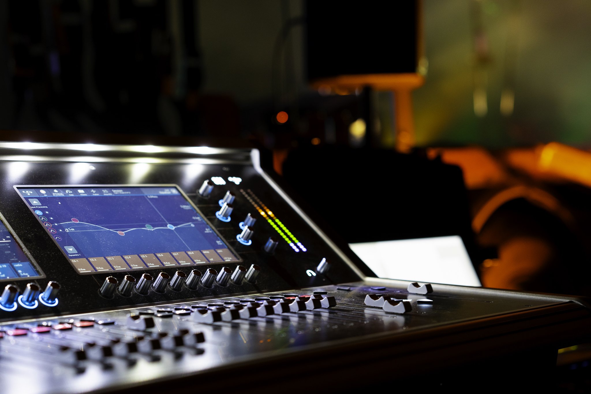 Close-up of a professional audio mixing console with illuminated sliders, knobs, and a digital display showing audio levels, in a dark, colorful environment likely a recording studio or nightclub.