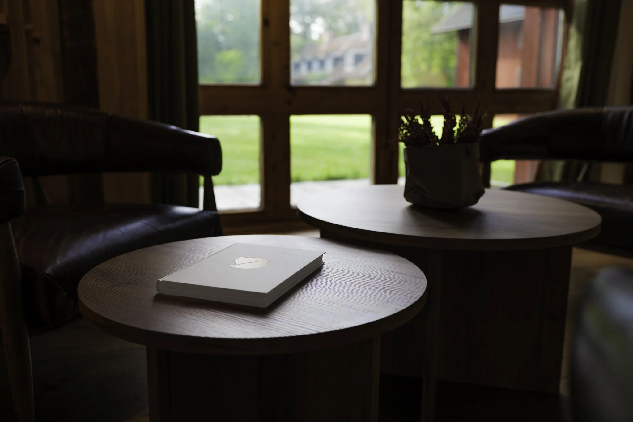 Wooden interior with two round tables, a book on one table, a potted plant on the other, leather chairs, large windows showing green outdoor landscape and houses.