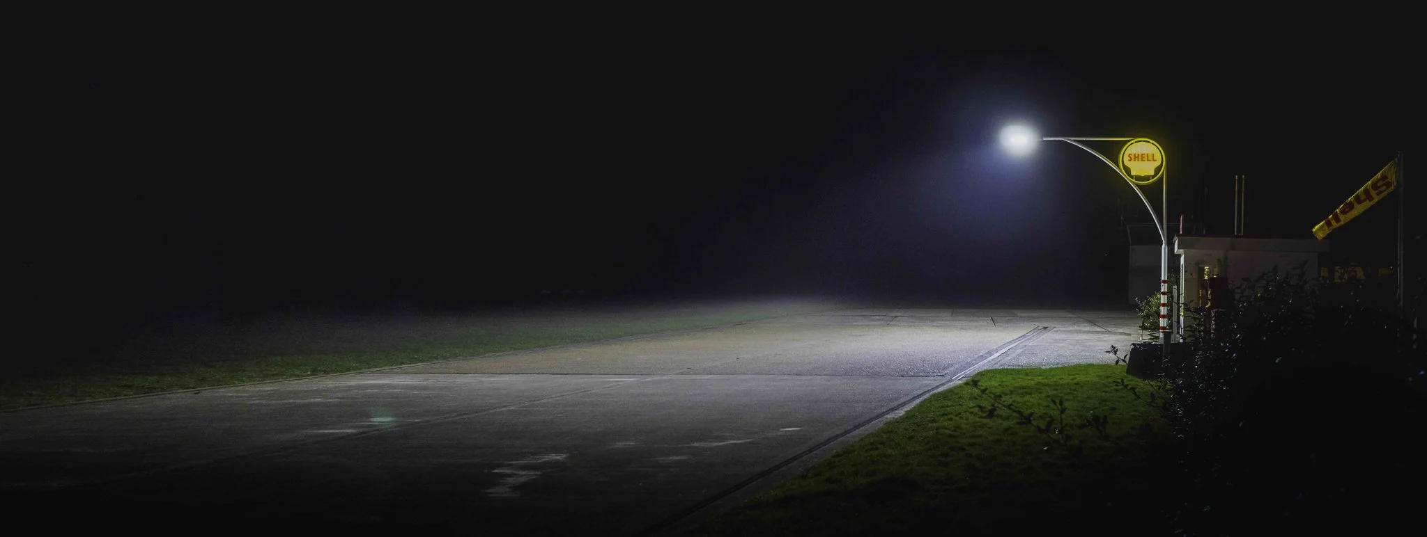Empty night street with illuminated Shell gas station sign and flag, dark sky, full moon, no cars or people visible.