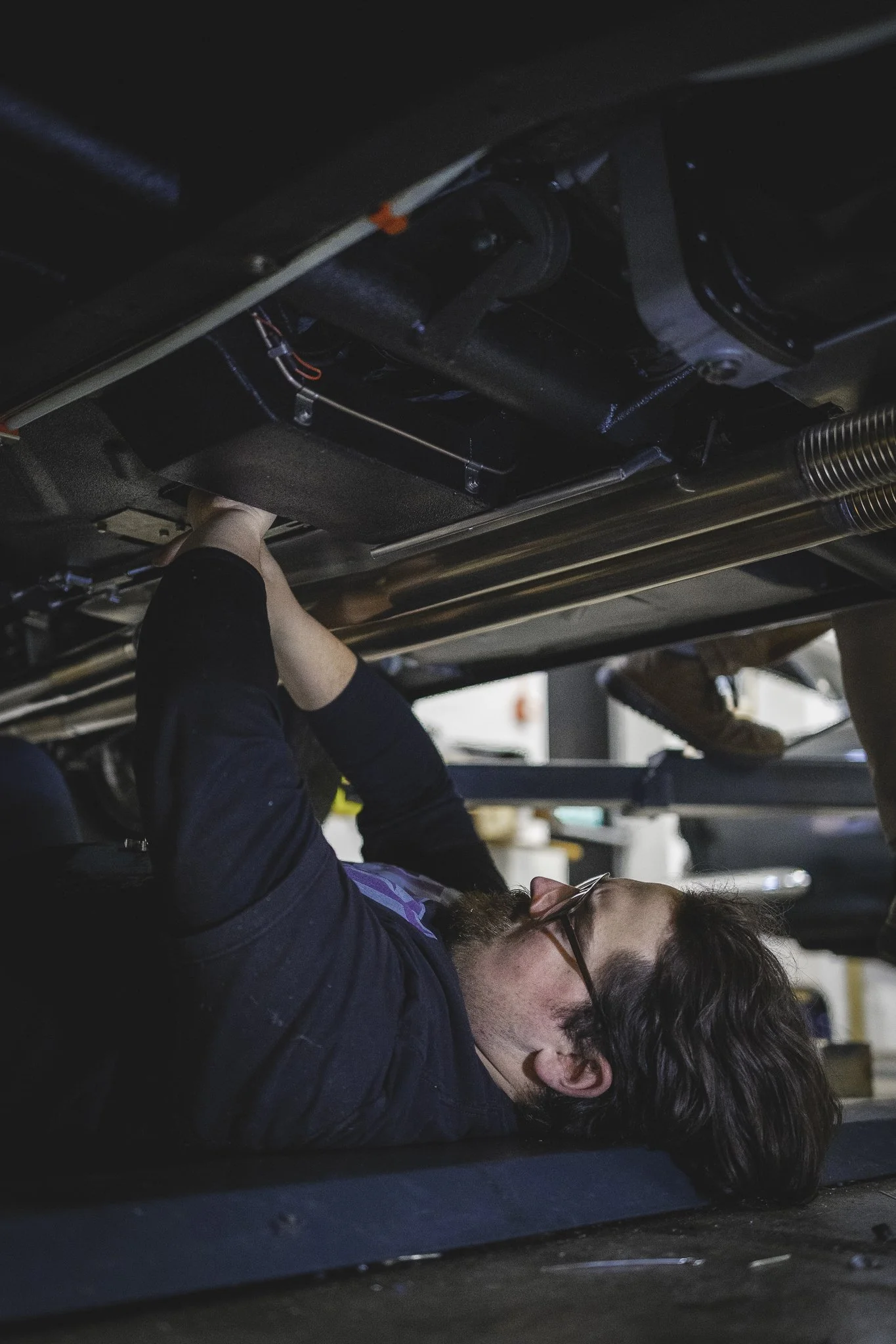 A person lying on their back under a vehicle, working on the underside, with tools and equipment visible in the background.