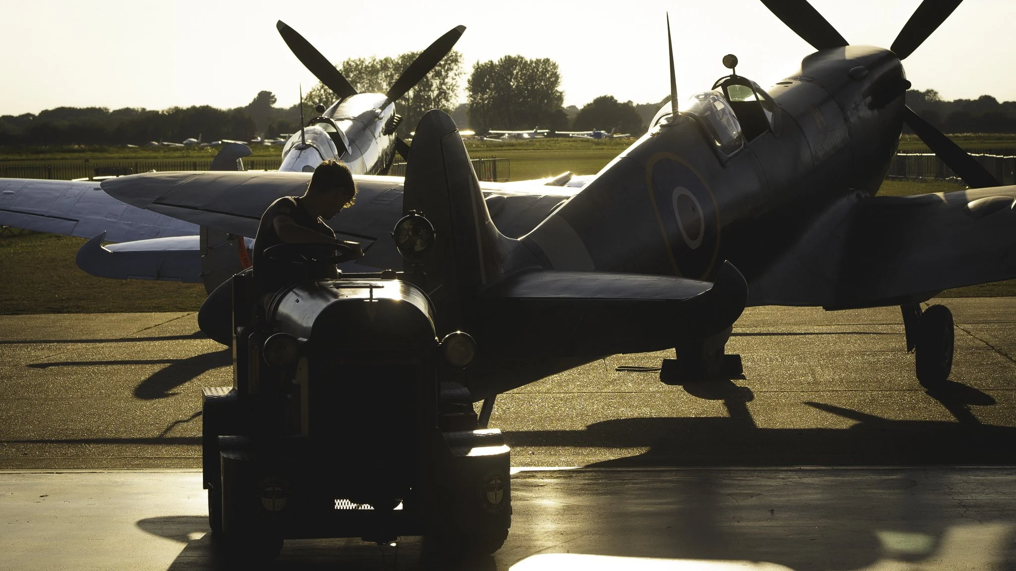 A person sitting on an aircraft tug on an airfield at sunset, with 2 spitfire fighter plane in the background.