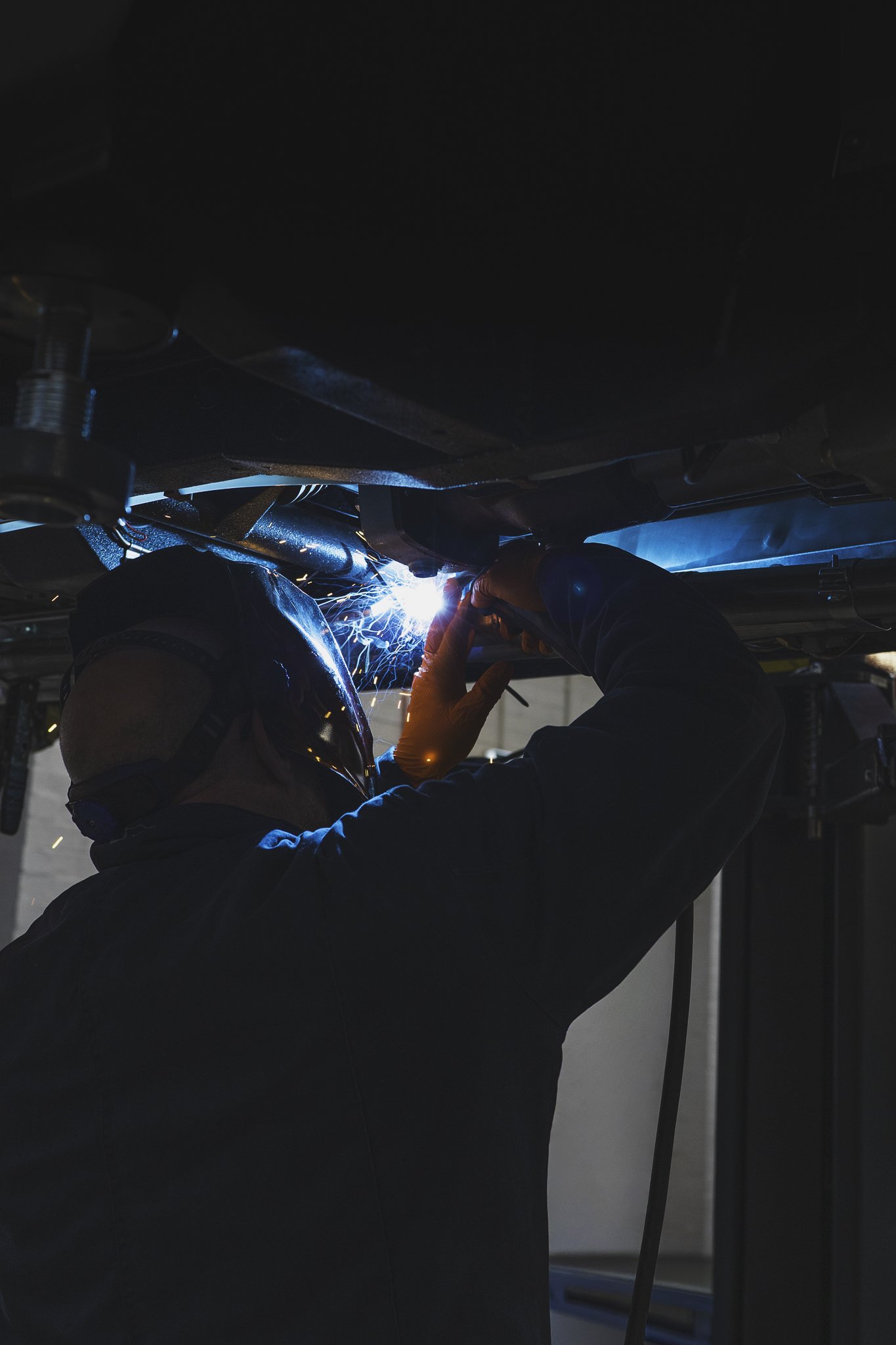 Welding technician wearing safety gear, welding under a vehicle in an industrial workshop.