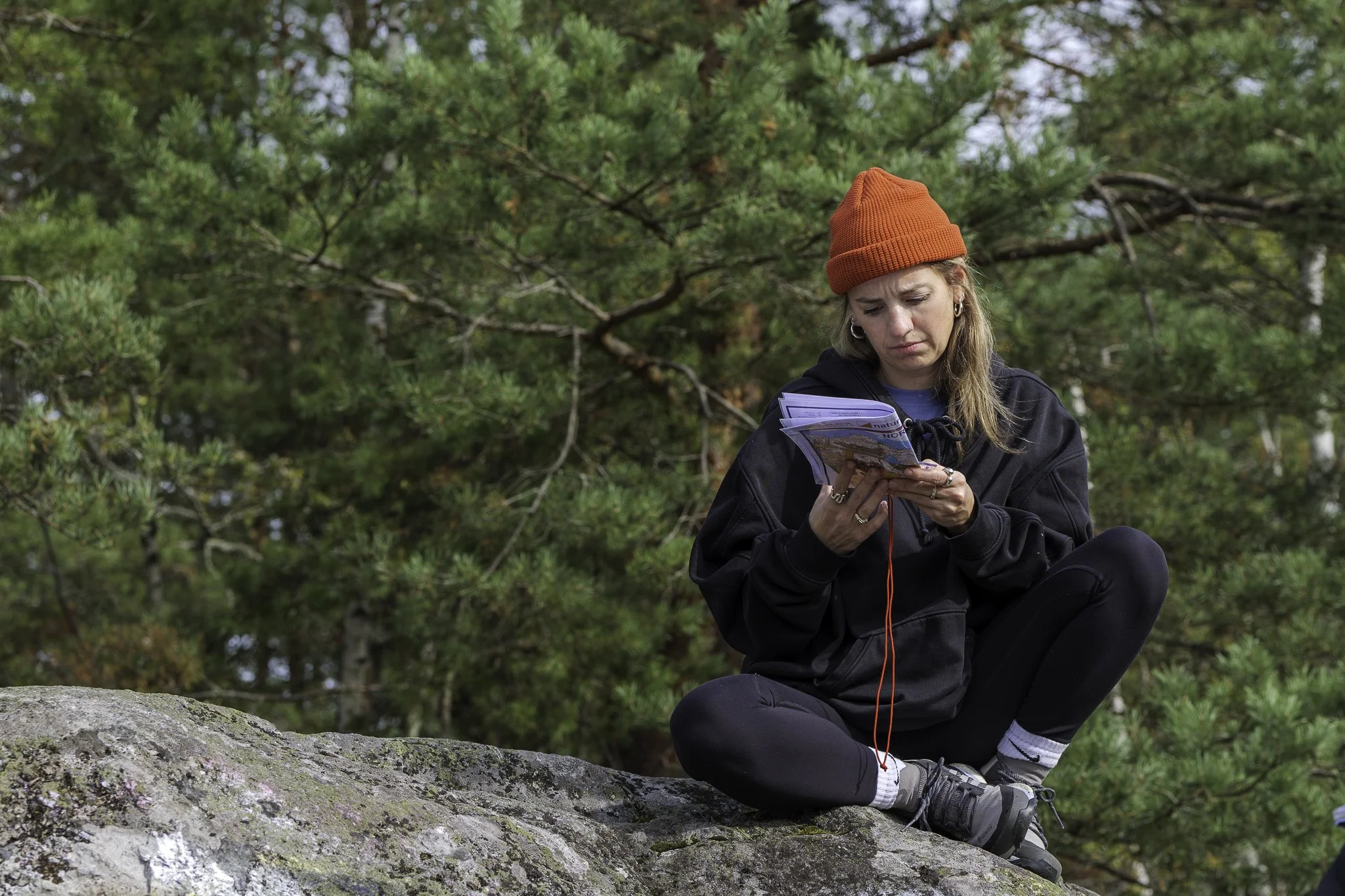 A young woman wearing a red beanie, black hoodie, and black leggings sits cross-legged on a large rock in a forest, looking at a map or brochure with a focused expression.