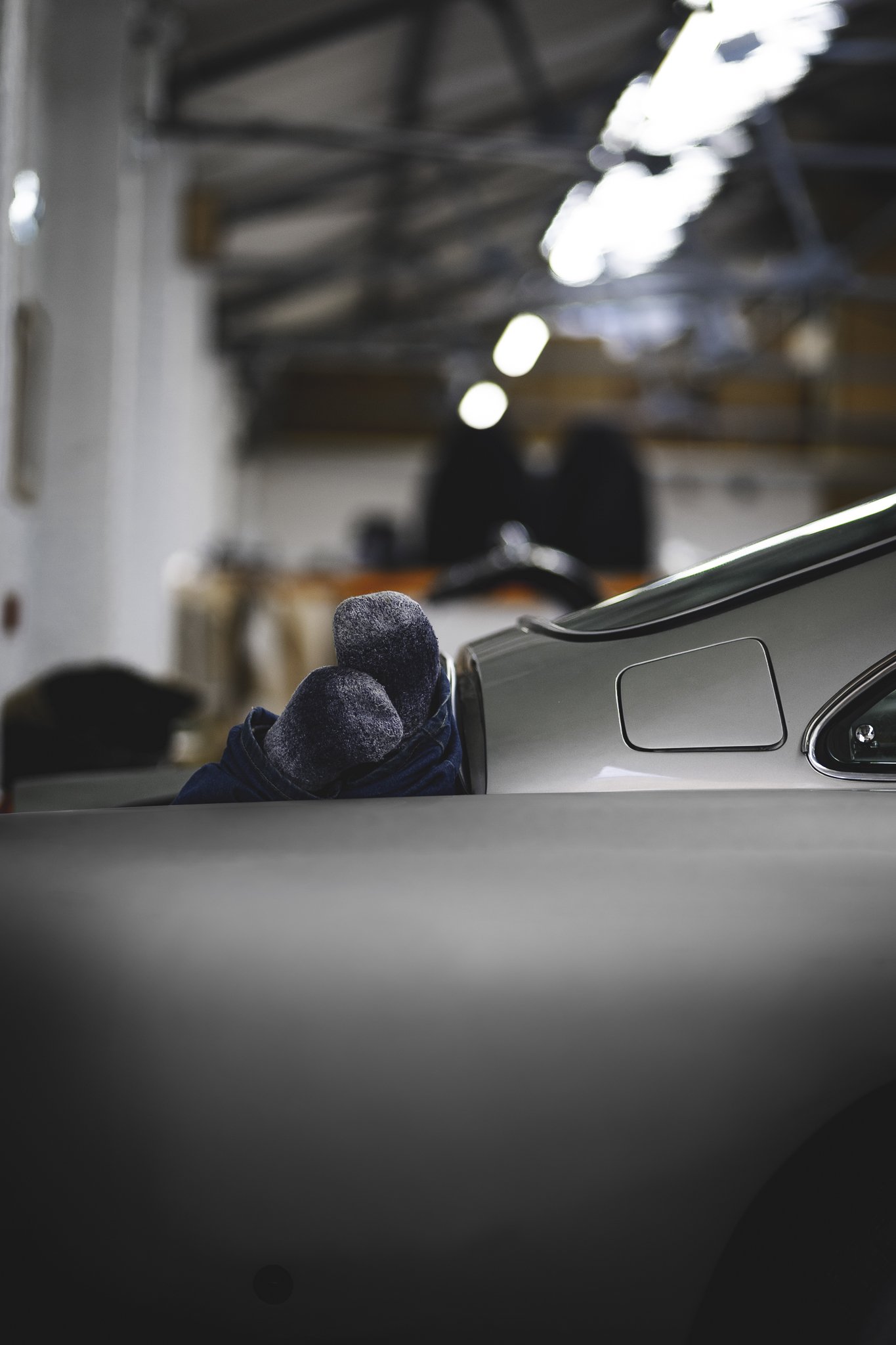 Close-up of a vintage gray car with a person wearing gray gloves resting against the side in a garage or warehouse, with blurred background.