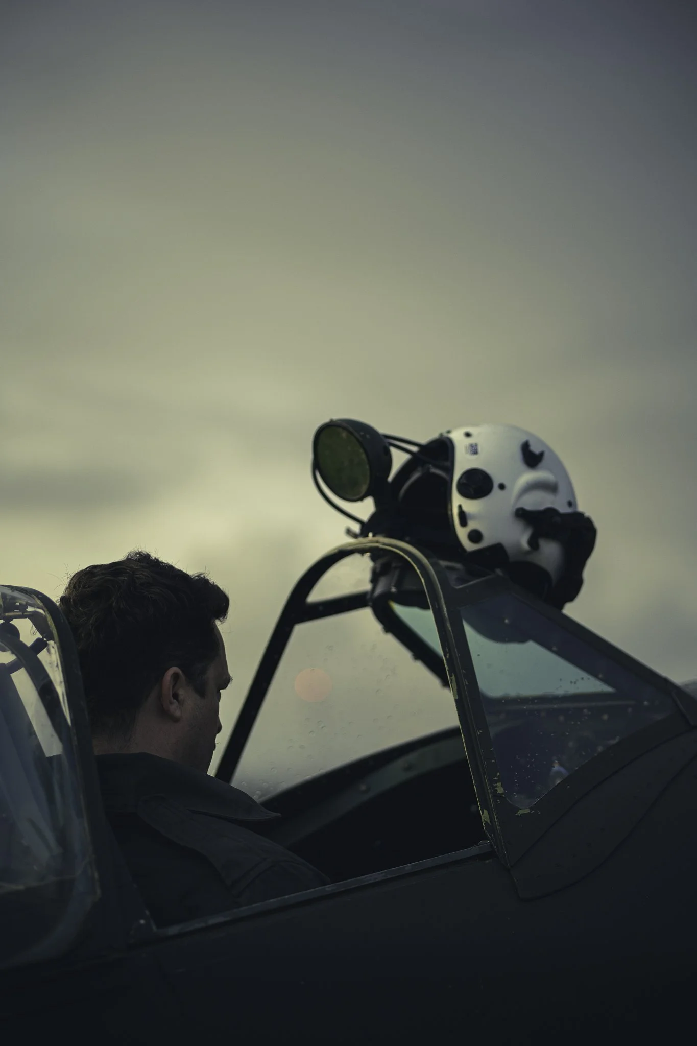 A man sitting in the cockpit of an aircraft with a helmet placed on the canopy frame.