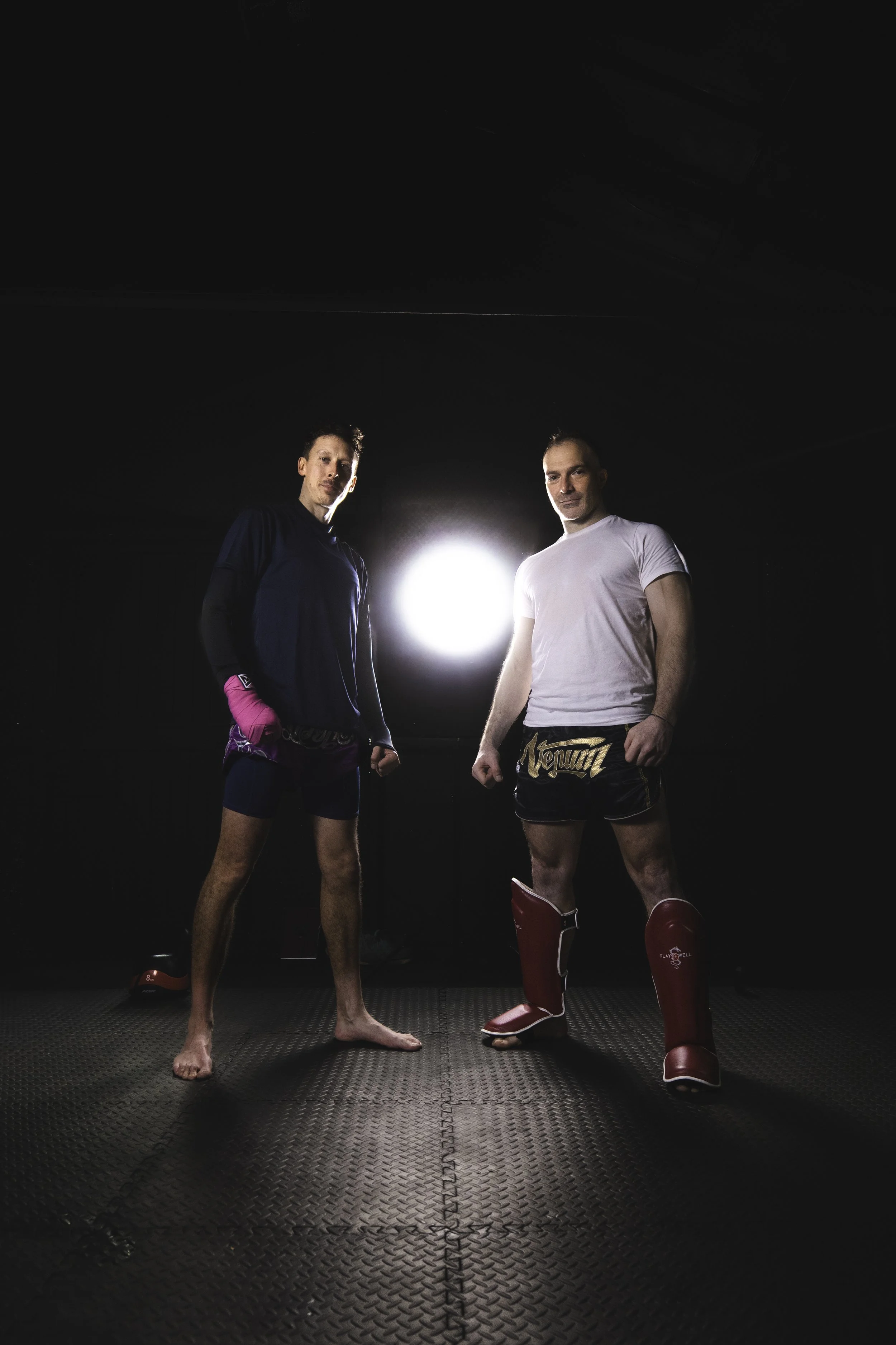 Two men in fighting gear standing on a black mat with a bright light behind them in a dark room.