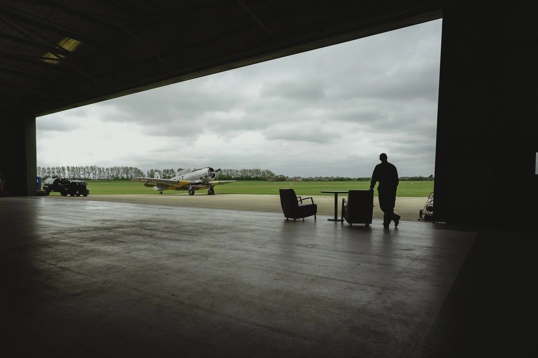 Silhouette of a person standing inside a hangar, with vintage fighter plane outside on the airport tarmac and chairs near a table.