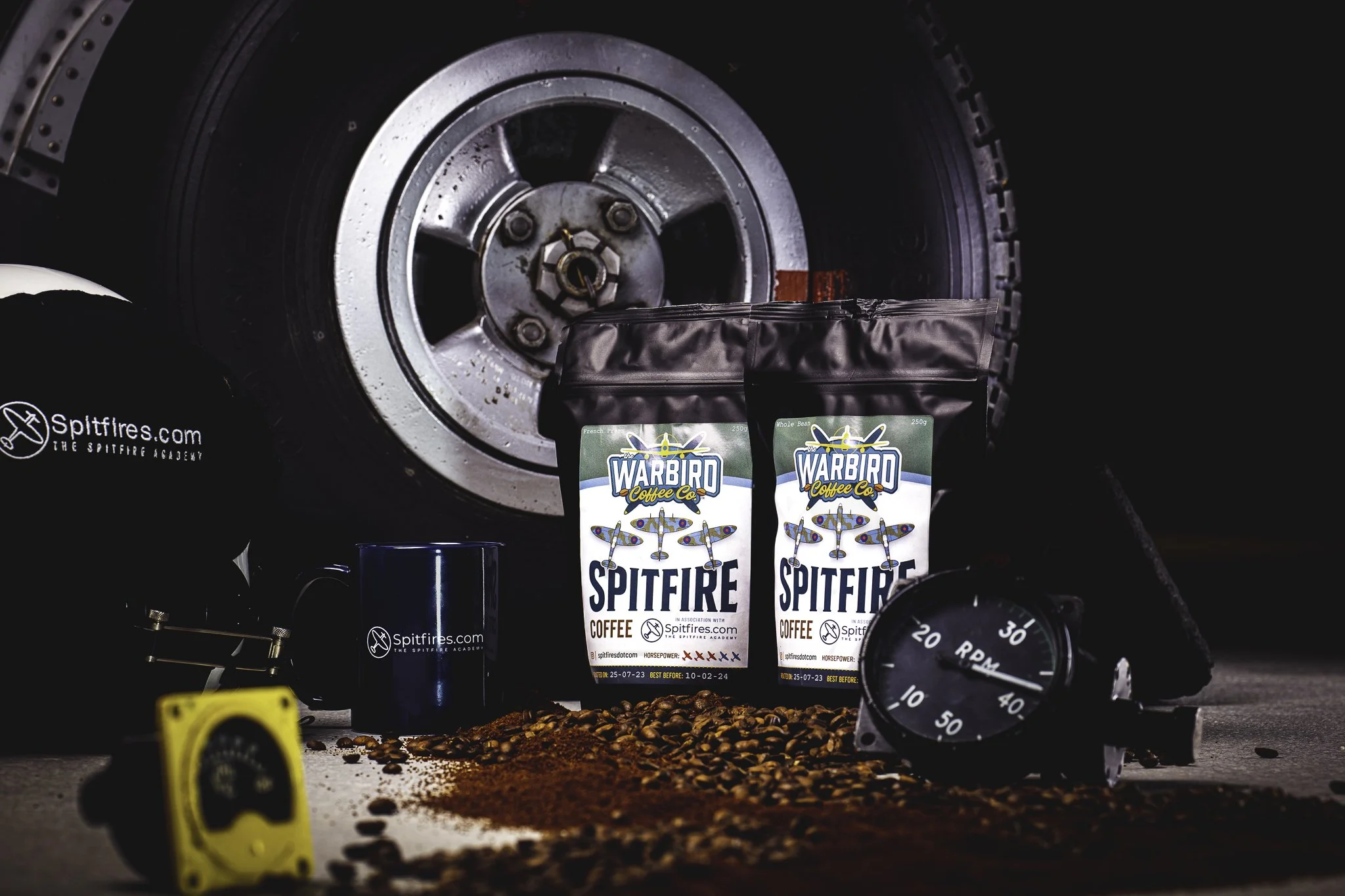Two packs of Warbird Spitfire coffee bags, a black helmet with white text, a blue coffee mug, a yellow measuring tape, coffee beans, ground coffee, a black watch, and a tire in the background, placed on a surface.