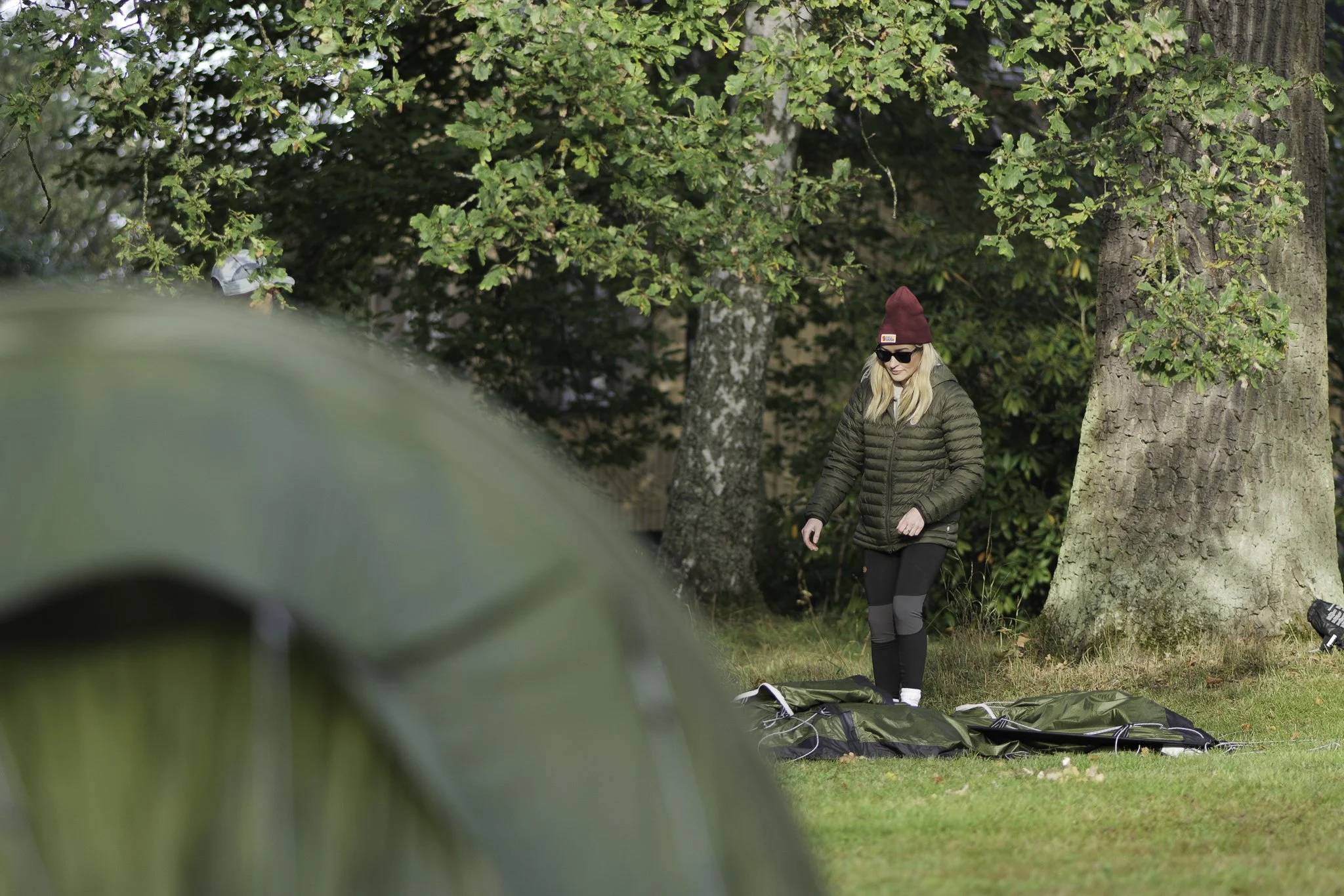 A woman with blonde hair, wearing sunglasses, a maroon beanie, a green puffer jacket, and black leggings, stands outdoors on a grassy area near trees, looking at camping gear laid out on the ground.