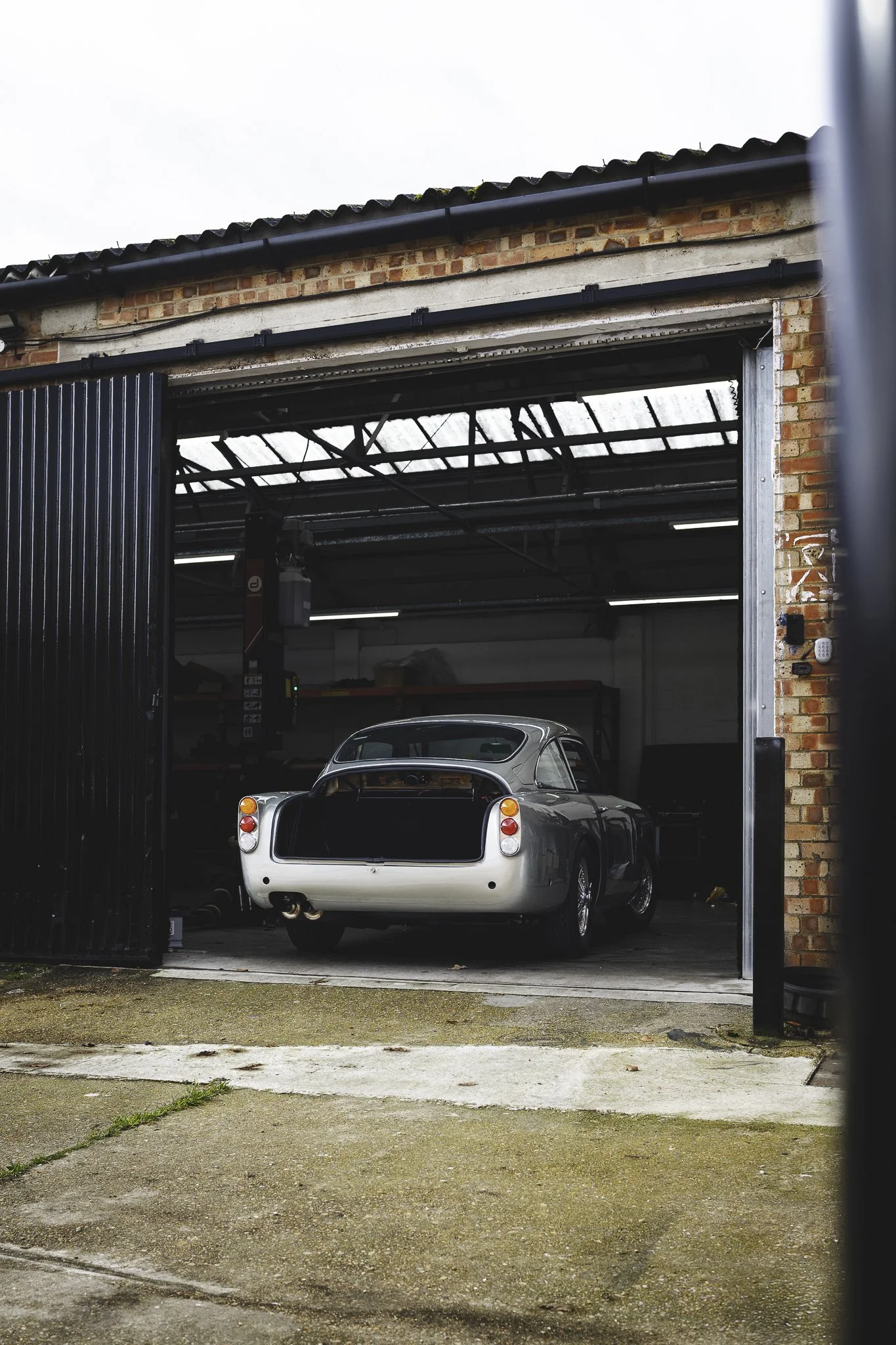 A vintage silver car parked inside a garage with open sliding doors, showing an industrial building with a brick exterior.