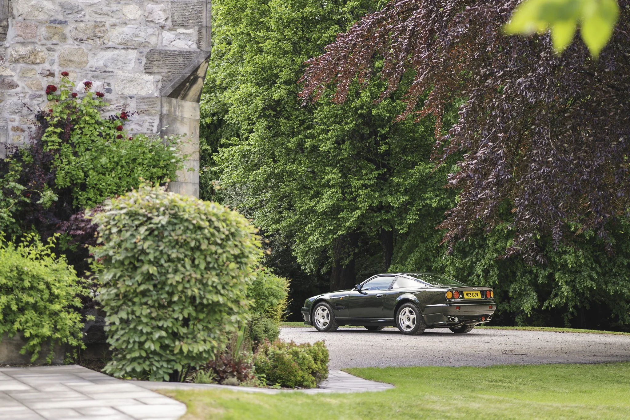 A black car parked on a driveway surrounded by lush green trees and vibrant bushes near a stone building.