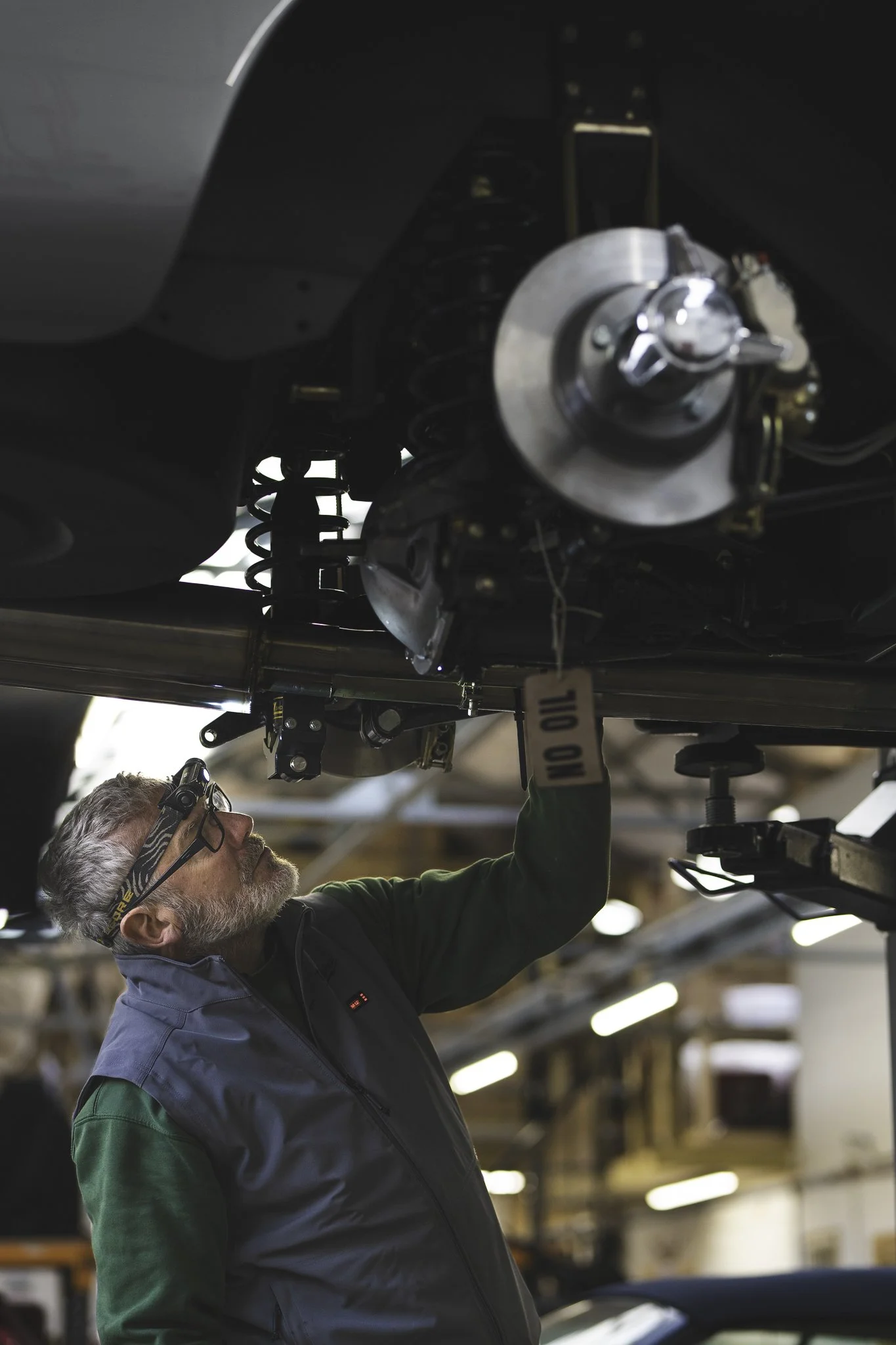 A man wearing glasses and a headlamp inspects the underside of a car, in a workshop.