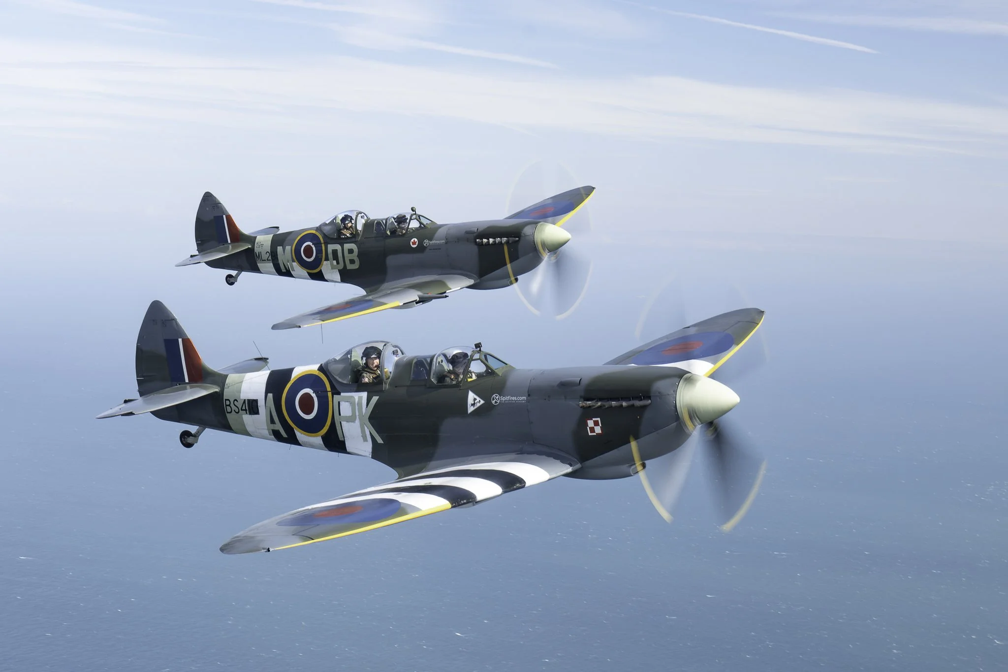 Two vintage military fighter planes flying in formation over the ocean, propellers spinning, with pilots visible in the cockpits.