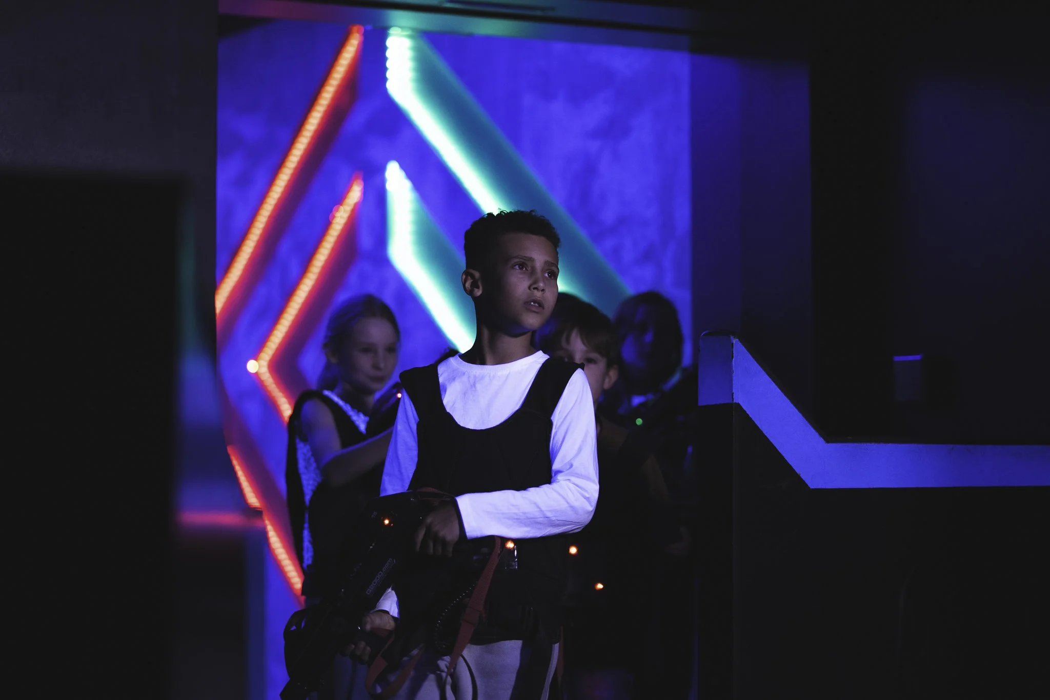 A group of children at a laser tag arena with colorful, bright neon lights in the background. One boy in the foreground is holding a laser tag gun and looking intently forward, while other children stand behind him.