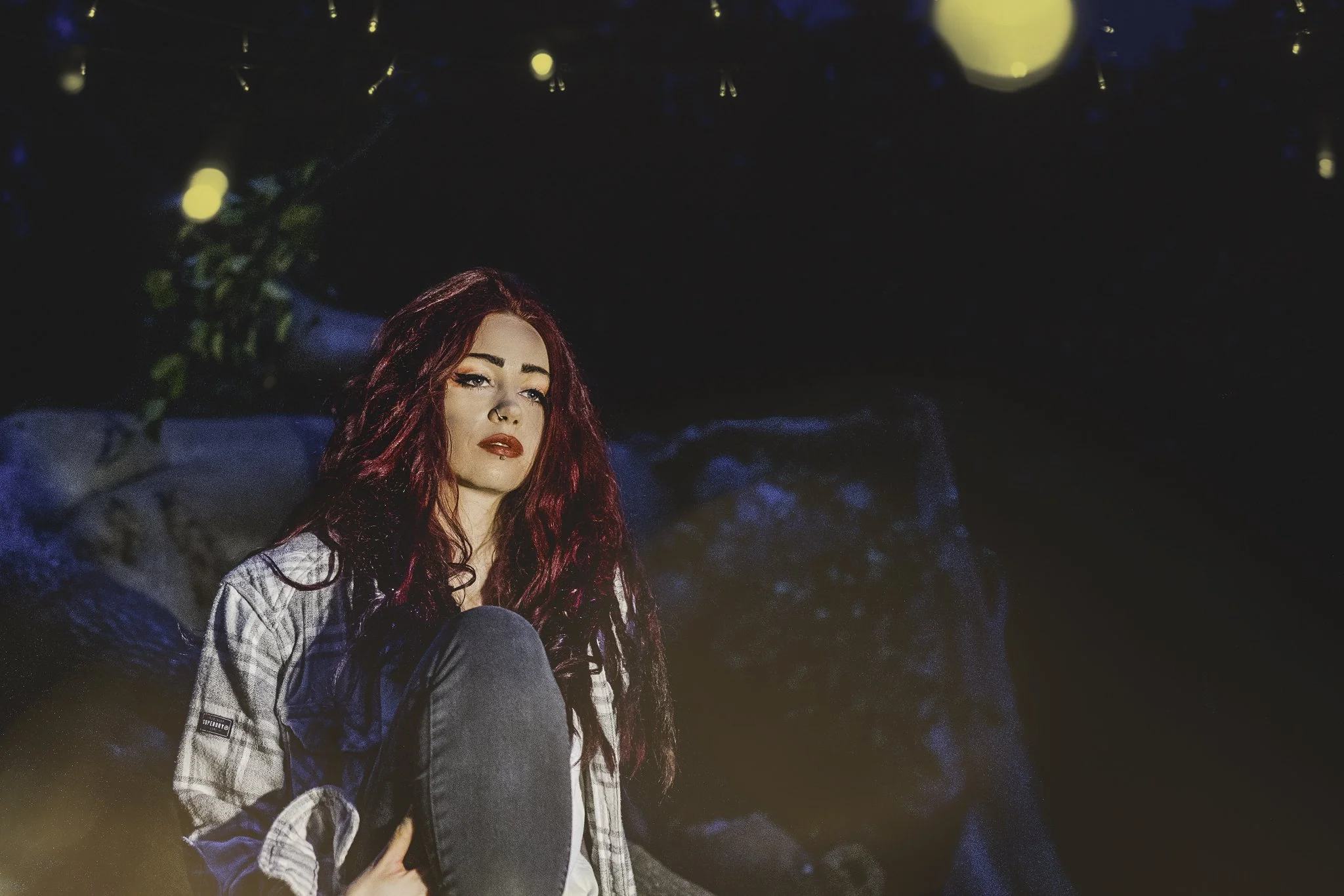 A woman with long red hair and makeup sitting outdoors at night with string lights overhead, wearing a plaid shirt and holding a hat.