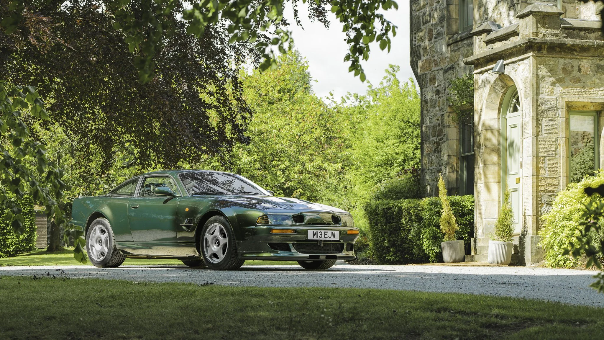 A green sports car parked on a driveway in front of a stone house surrounded by trees and plants.