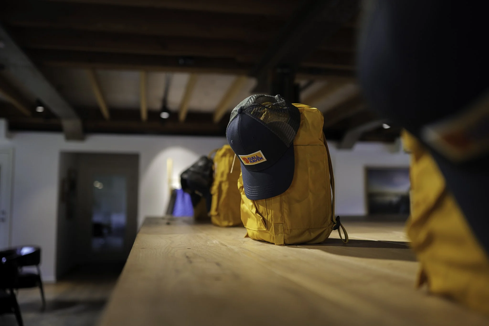 Yellow backpacks with a black Billabong cap hanging on one in a row on a wooden counter in a room with a wooden ceiling and a door in the background.
