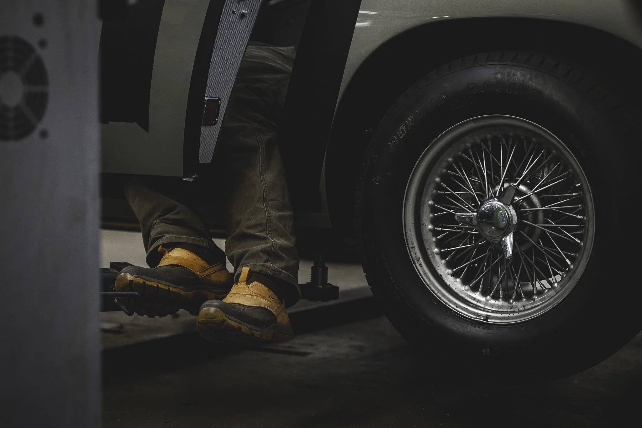 Close-up of a person wearing tan work boots and brown pants, sitting in a car with wheel and wire spoke rim visible, appearing to work on the vehicle at a garage.