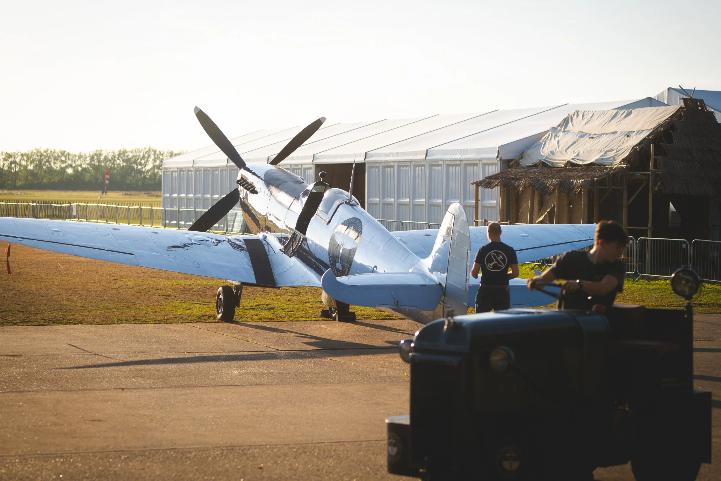 A vintage silver aircraft parked on an airfield in front of hangars, with two people standing nearby, at sunset.