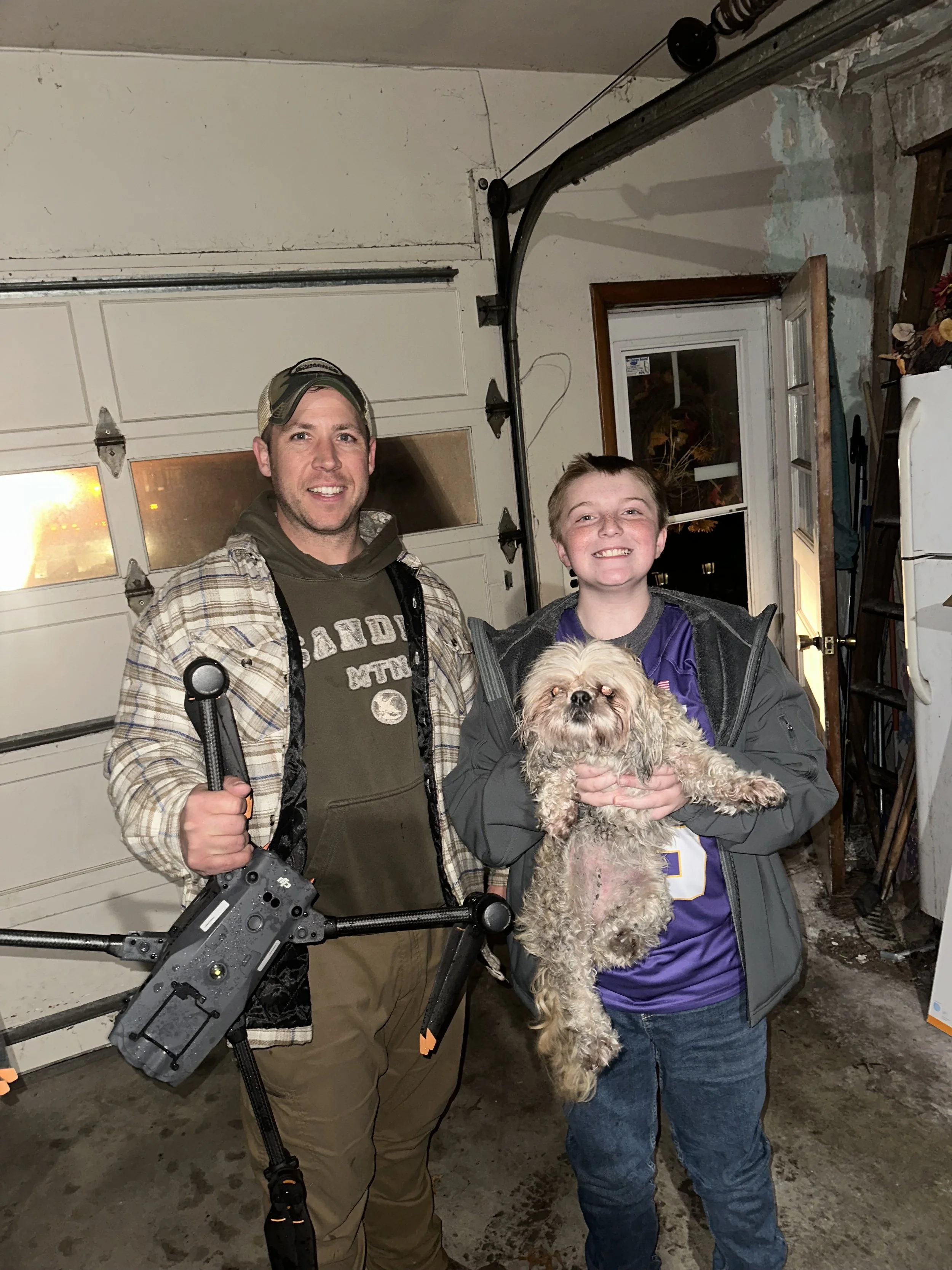 A man and a child holding a dog inside a garage. One man holds a drone frame. The boy is smiling and the dog is looking at the camera.