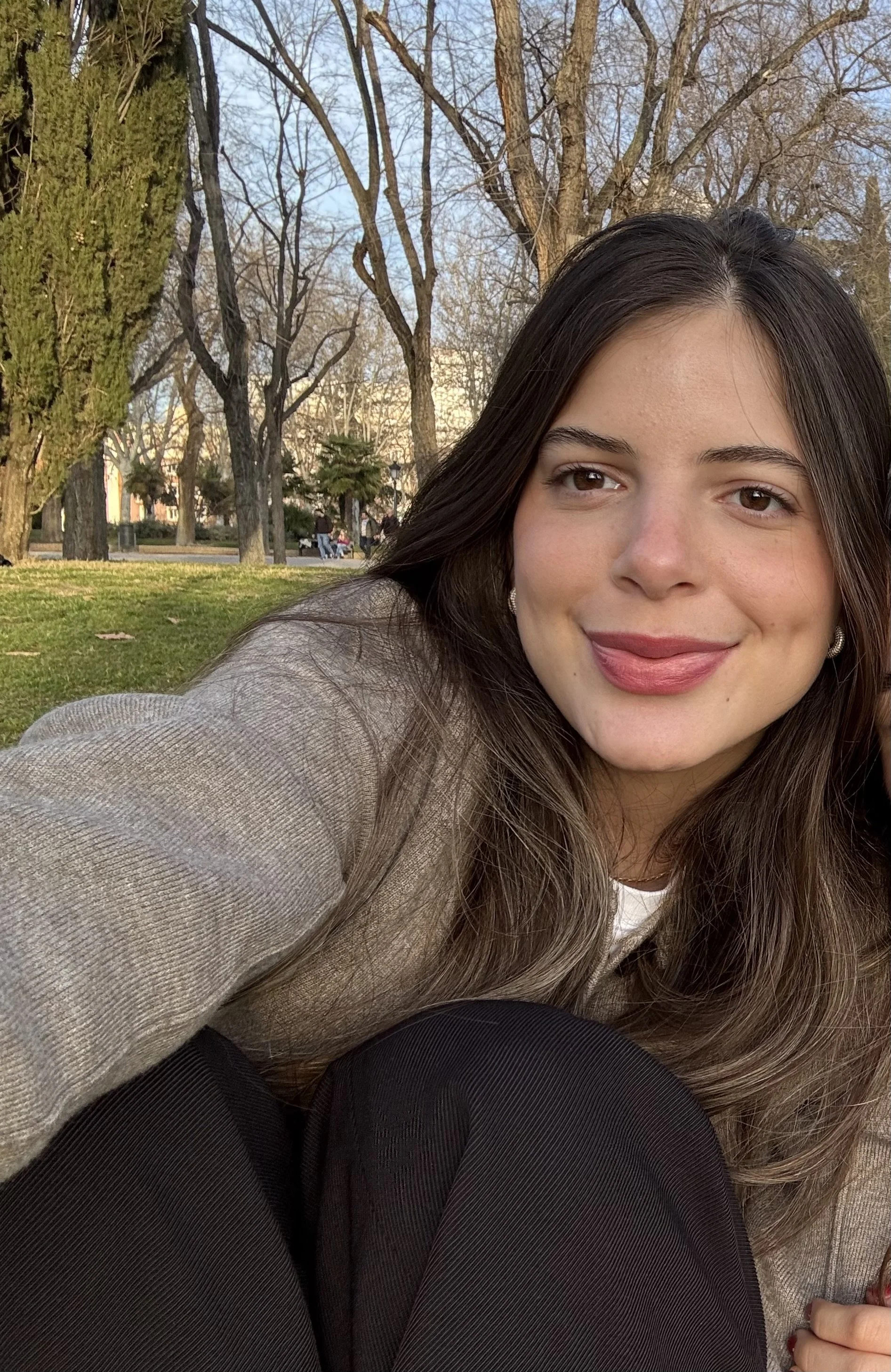 A young woman with long brown hair, wearing a beige blazer and black pants, smiling while sitting in a park with trees and a few people in the background.