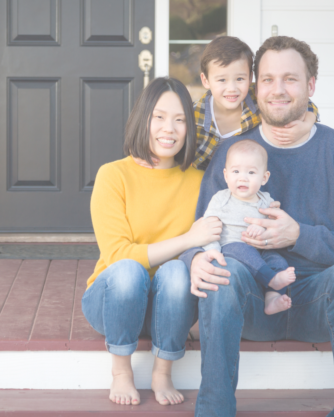 A family of four sitting on their front porch in front of a black door, smiling at the camera. The mother is wearing a yellow sweater and blue jeans, the father is in a blue sweater, the young boy on his lap is wearing a yellow and blue plaid shirt, and the baby is in a gray onesie.