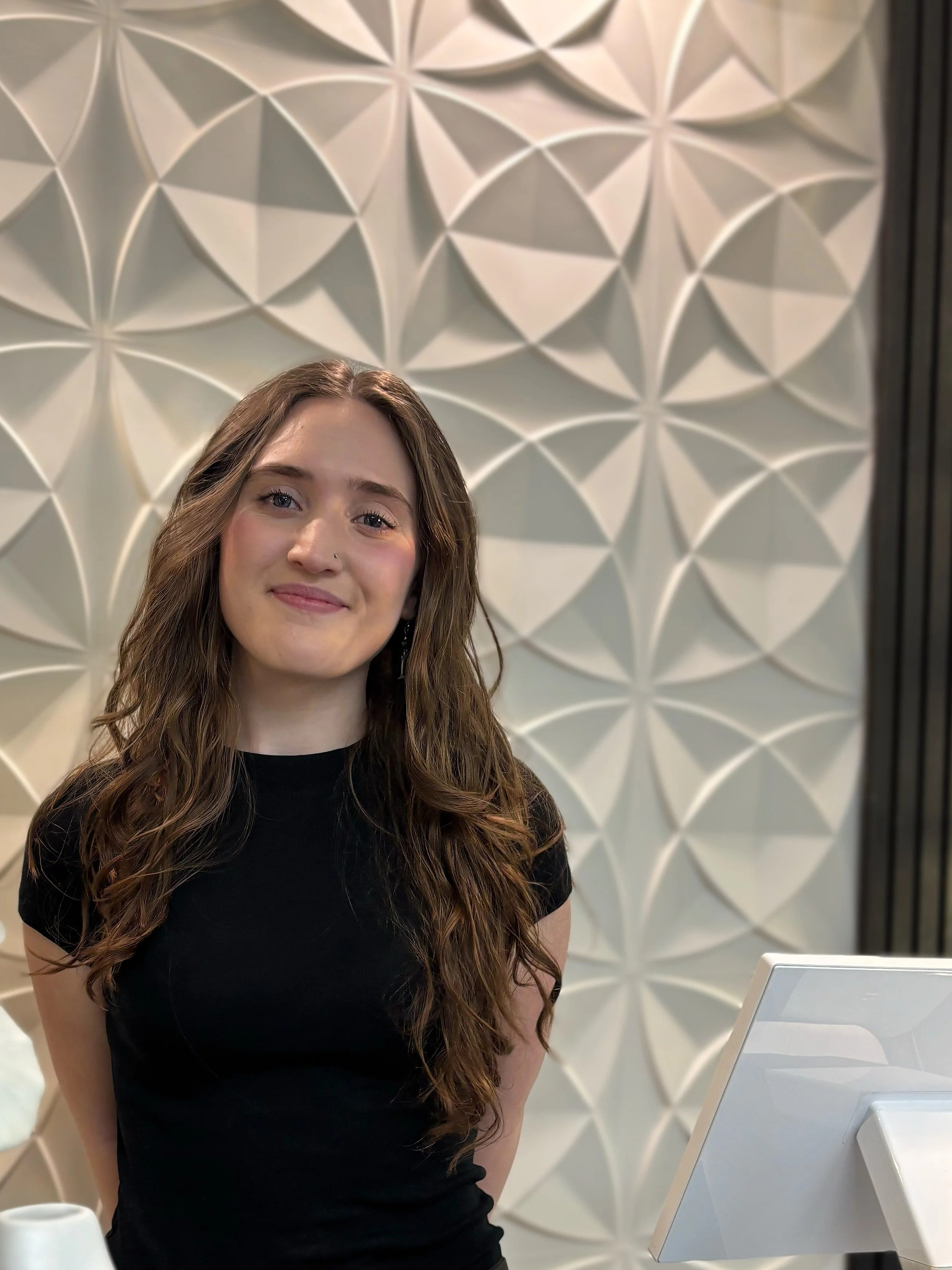 A woman with long brown hair is standing in front of geometric wall art and smiling.