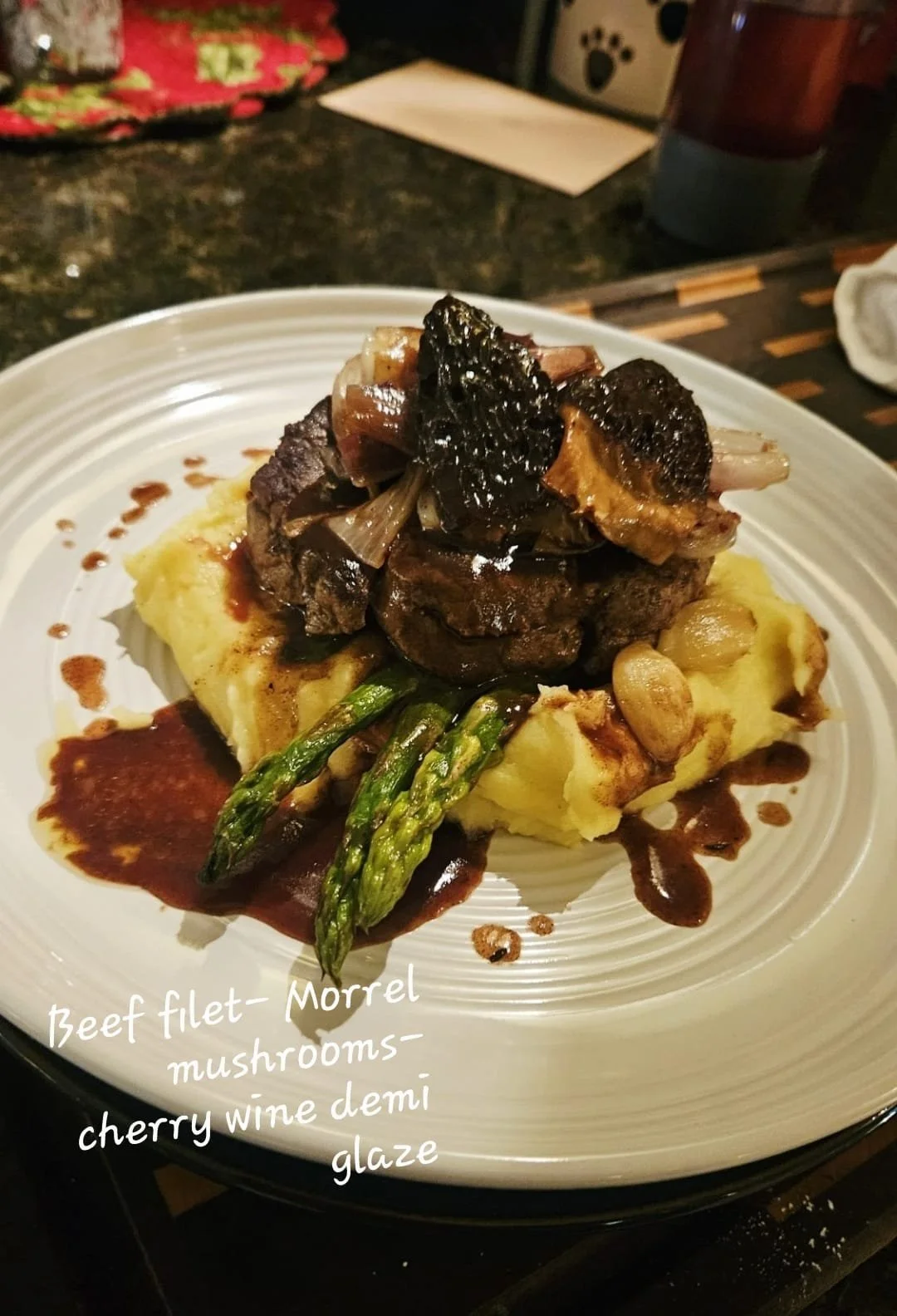 A plate of beef fillet topped with mushrooms, cherry wine demi-glaze, and garnished with asparagus and garlic, served on a bed of mashed potatoes.