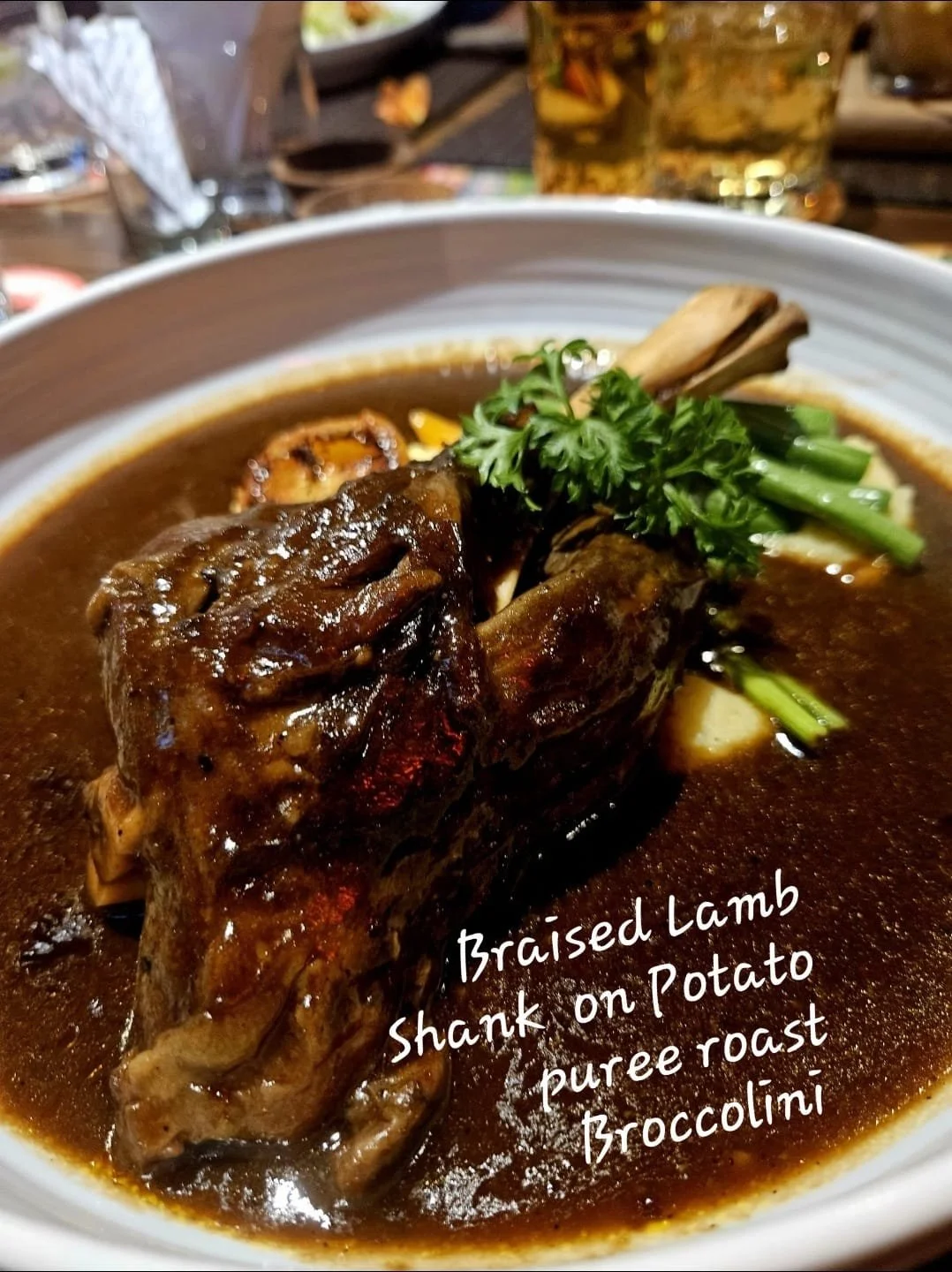 A dish of braised lamb shank on potato puree and broccoli, served in a bowl with a rich sauce, garnished with herbs.