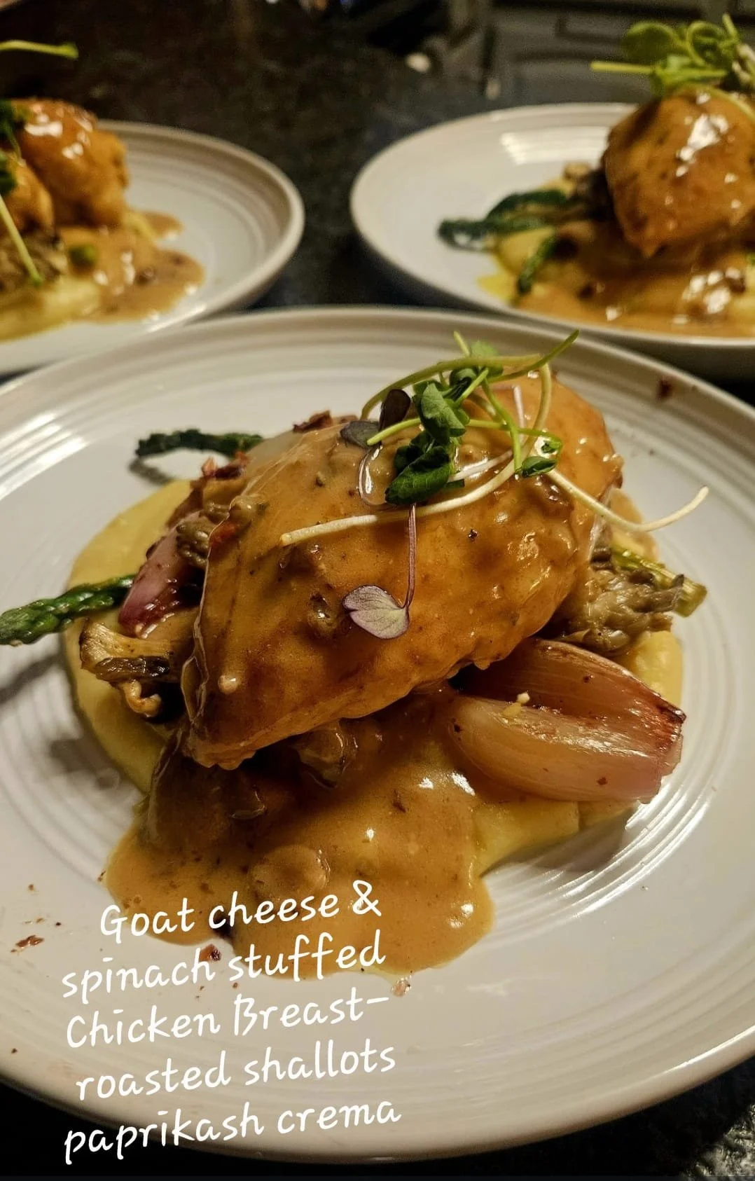 A plated dish of roasted chicken breast topped with goat cheese and microgreens, served with spinach-stuffed roasted shallots and a creamy paprika crema sauce, on a white plate.