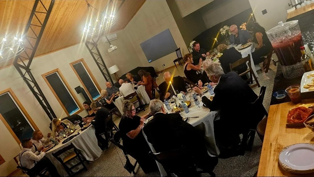 People attending a formal dinner party in a decorated room with string lights, tables set with white tablecloths, wine bottles, and food.