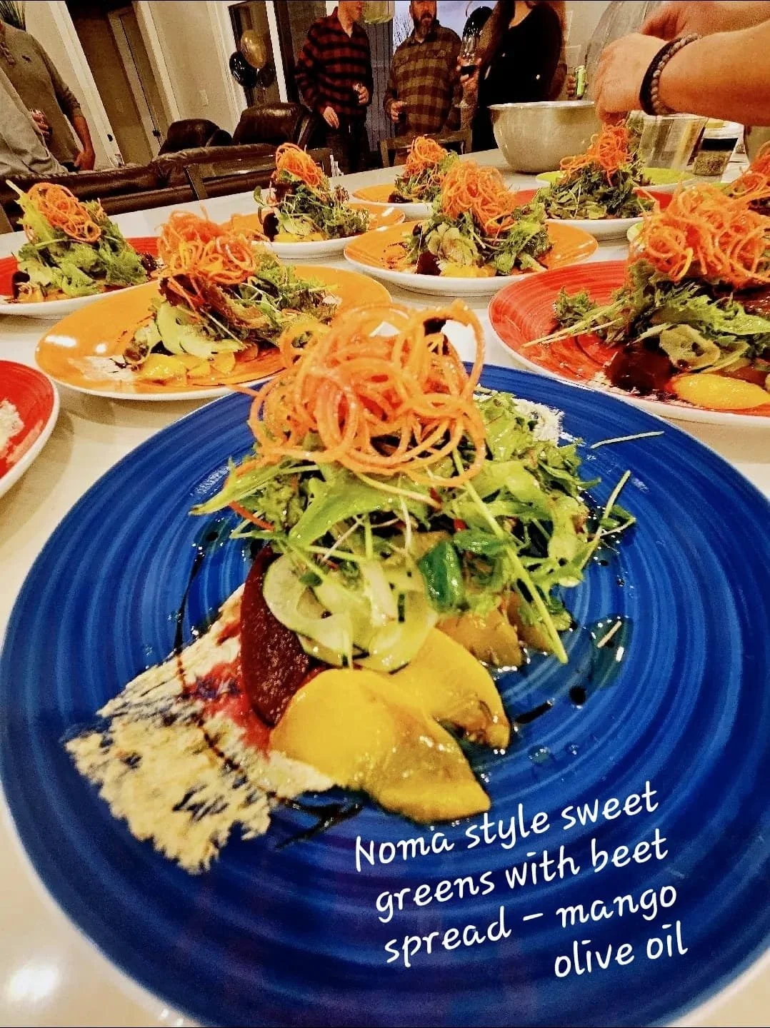 A colorful plated salad topped with spiralized carrots and mixed greens, with a description mentioning 'Noma style sweet greens with beet spread, mango, and olive oil' written on the plate. Several similar salads are on the table, and people are gath