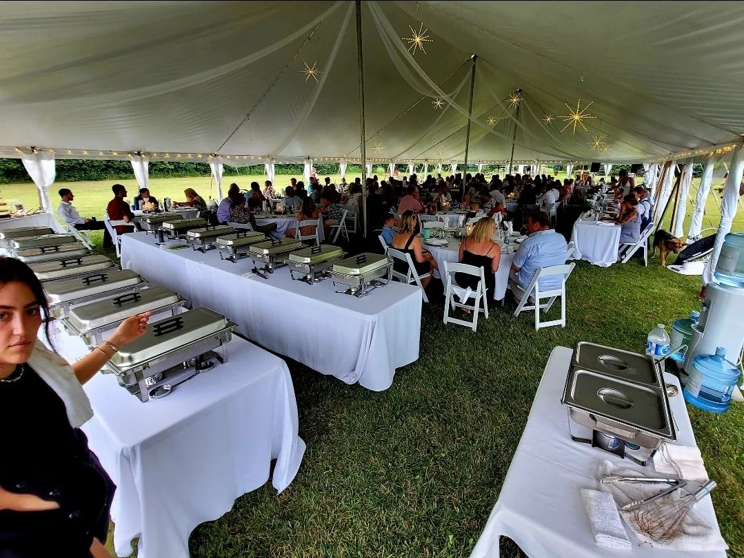 A large outdoor event tent with string lights and star-shaped decorations, filled with guests sitting at round tables and a buffet setup with chafing dishes on tables covered with white tablecloths.