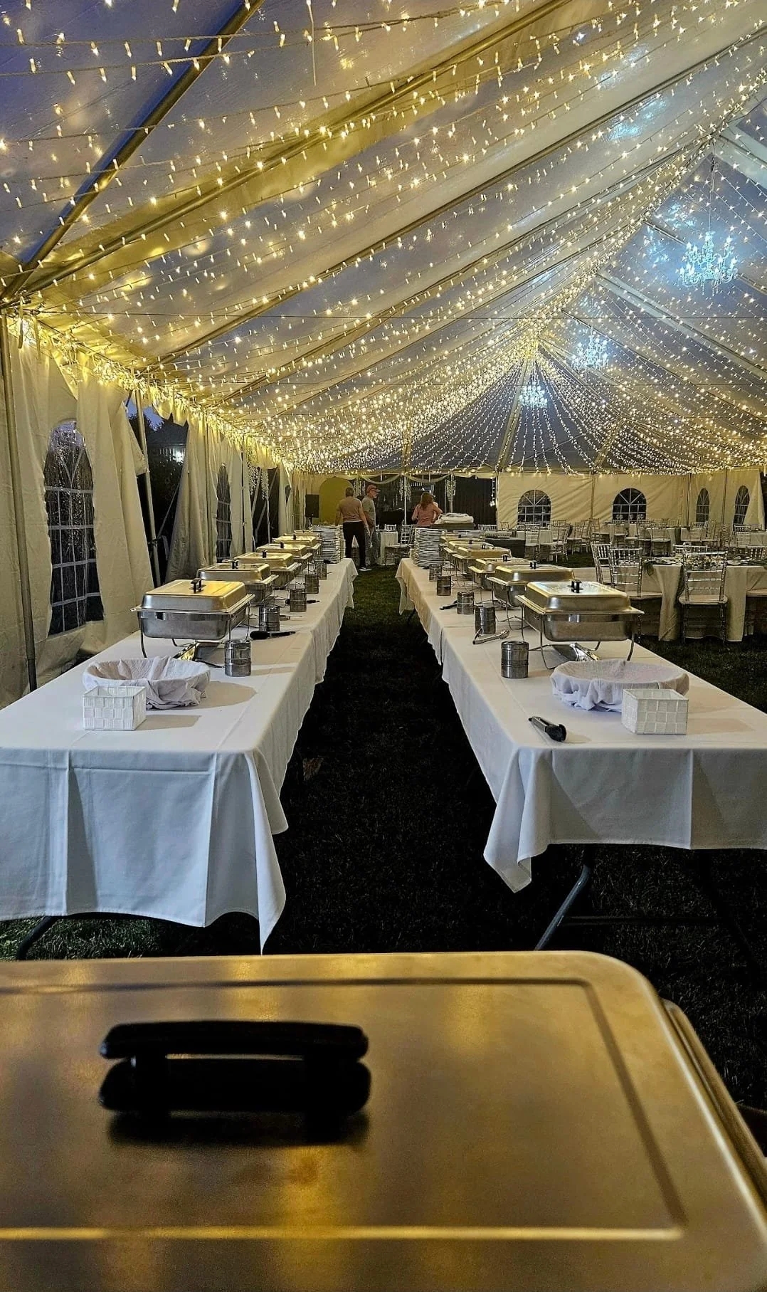 Empty banquet tent decorated with string lights, set up with long tables covered with white tablecloths, each holding chafing dishes, glasses, and small baskets.