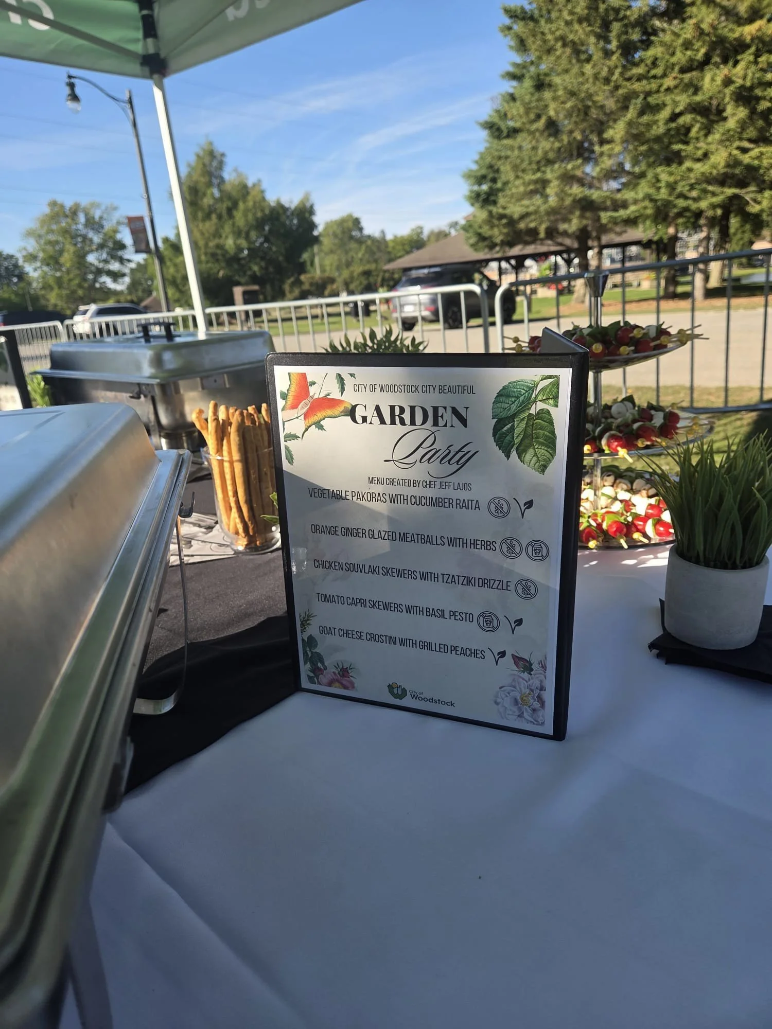 A food menu at an outdoor garden party in Woodstock, with a list of dishes including vegetable pakoras, cucumber raita, ginger glazed meatballs, chicken souvlaki skewers, tomato capri skewers, and goat cheese crostini. The background shows an outdoor