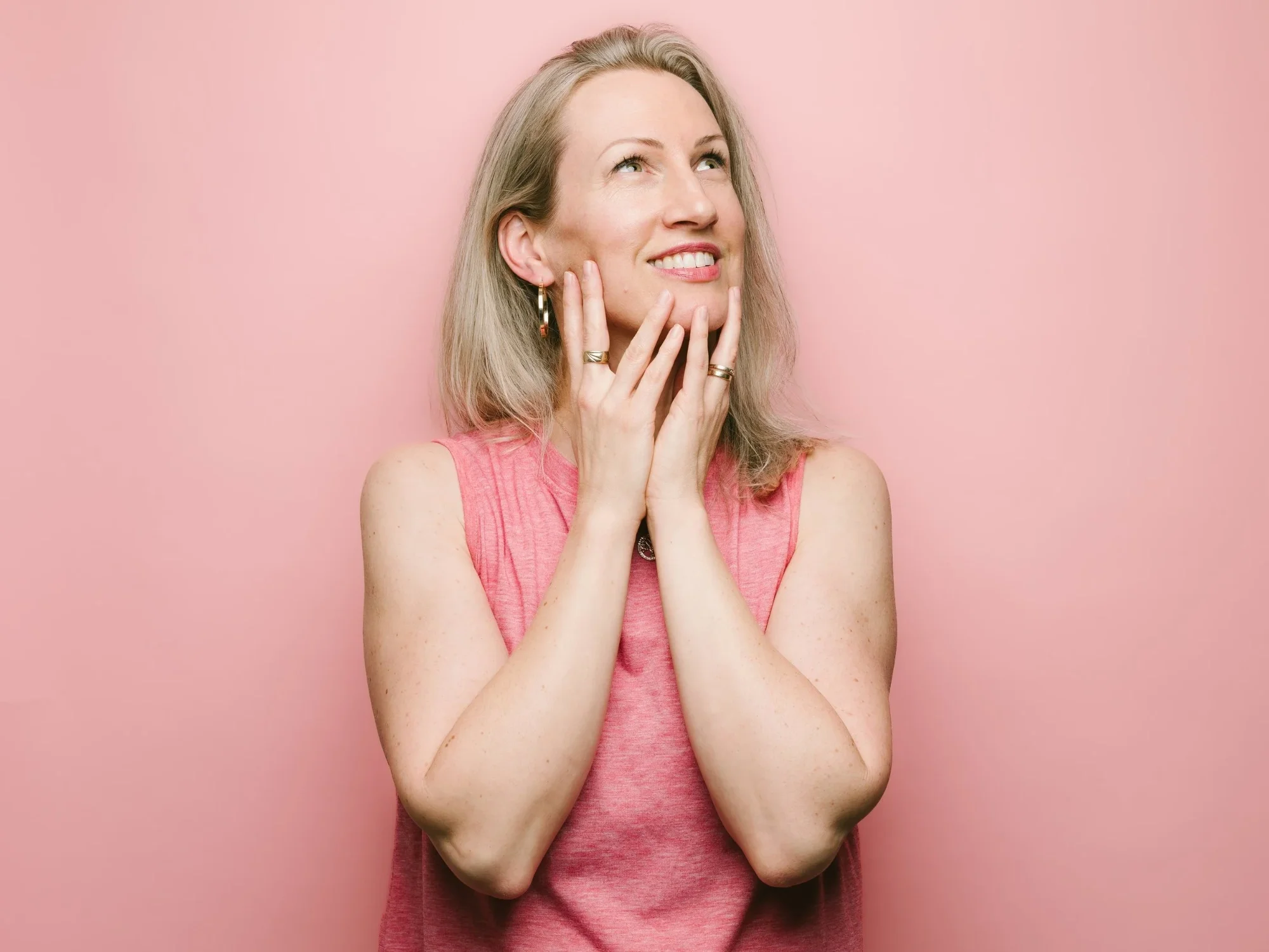 A woman with blonde hair and blue eyes, wearing hoop earrings and a pink sleeveless top, is smiling and looking upwards against a pink background.