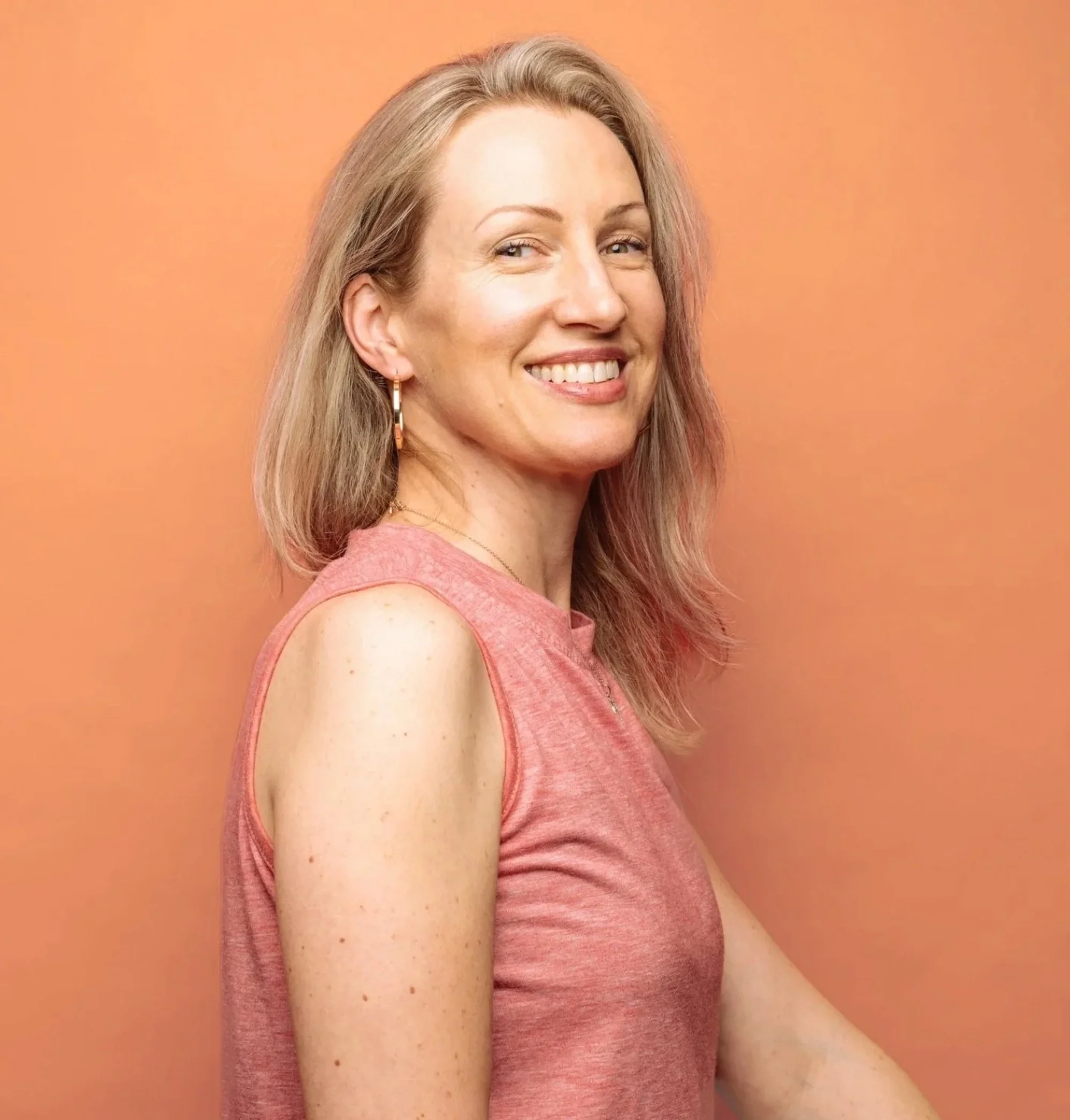 Close-up of a woman with shoulder-length blonde hair, green eyes, and fair skin, smiling, wearing a pink sleeveless top and gold earrings, standing against a peach-colored background.