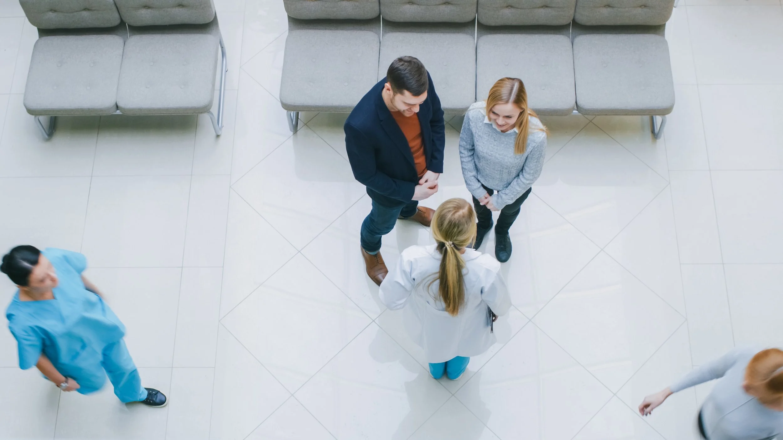 Three people, two standing and one sitting, talking in a waiting area of a hospital or clinic, with medical staff members nearby.