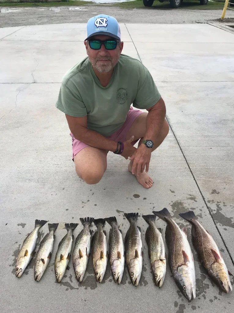 A man squatting beside a row of freshly caught fish spread on the ground, wearing sunglasses, a blue baseball cap with the North Carolina Tar Heels logo, a green t-shirt, pink shorts, and a watch, with a parking lot and a vehicle in the background.