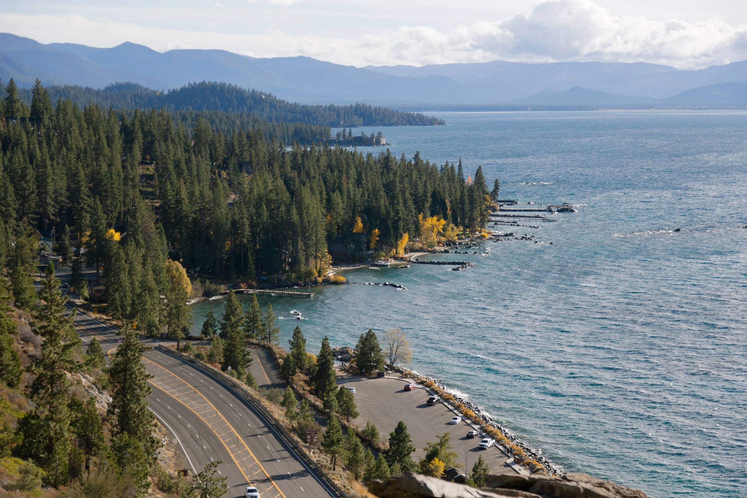 A scenic view of a coastal highway winding along a forested shoreline with a parking lot and the ocean beyond, mountains in the distance.