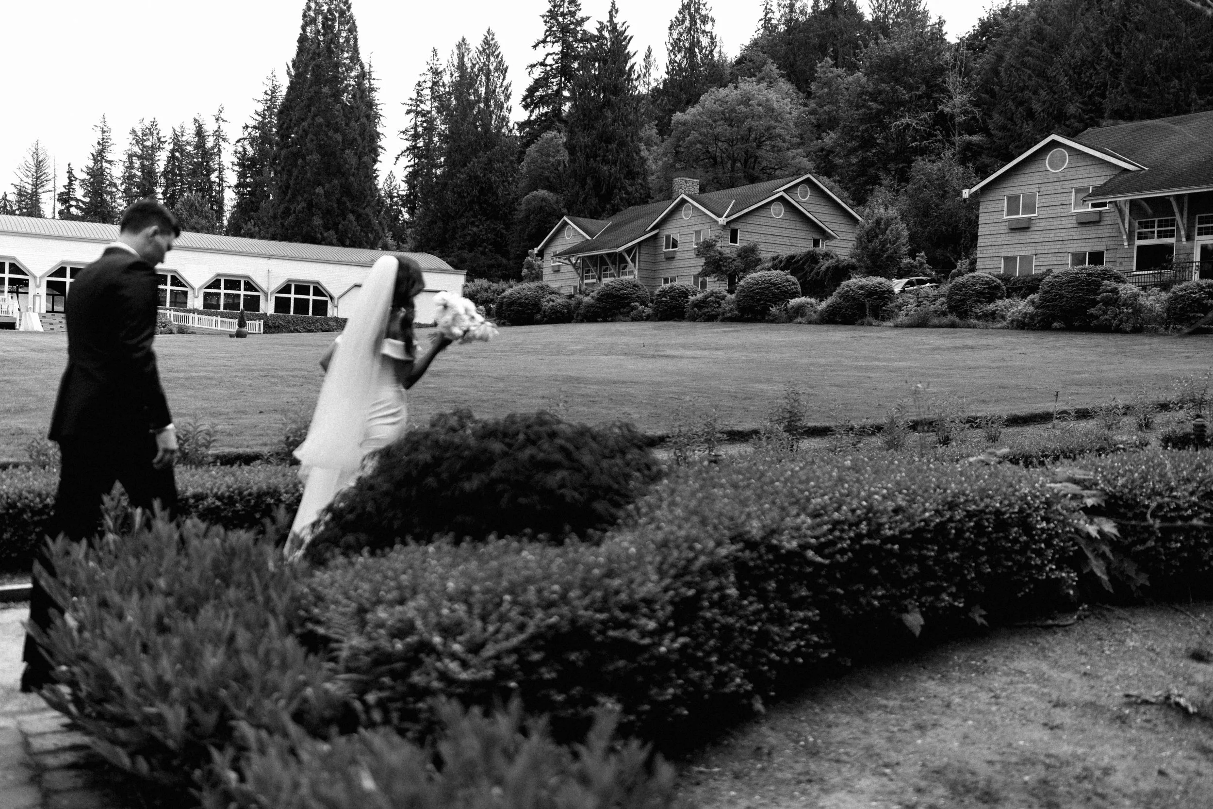 A black and white photo of a groom and bride walking outdoors on a grassy area, with the bride holding a bouquet.