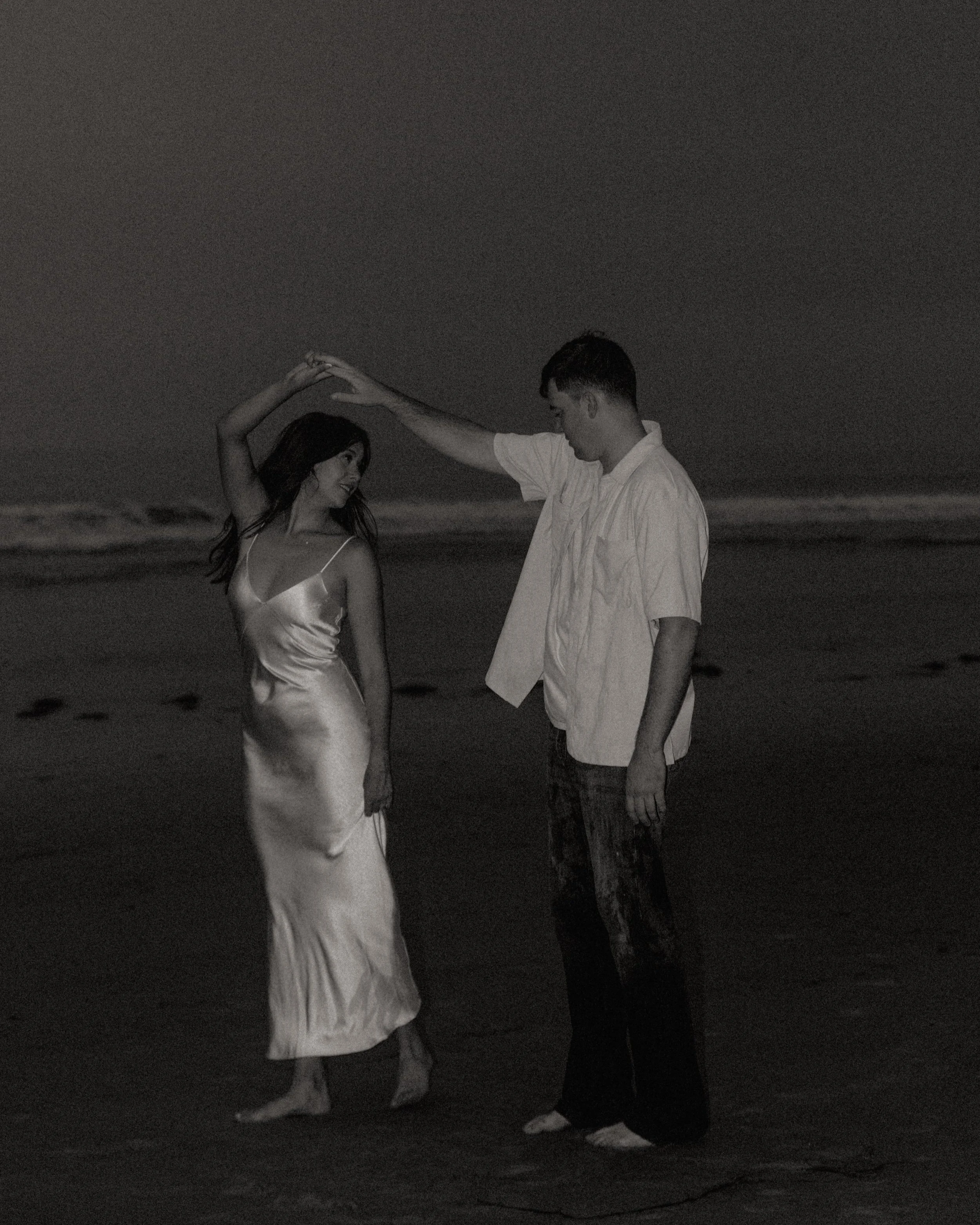 A black and white photo of a couple dancing on the beach at night, with the ocean in the background. The woman is wearing a satin dress, and the man is dressed casually in a short-sleeved shirt and jeans.