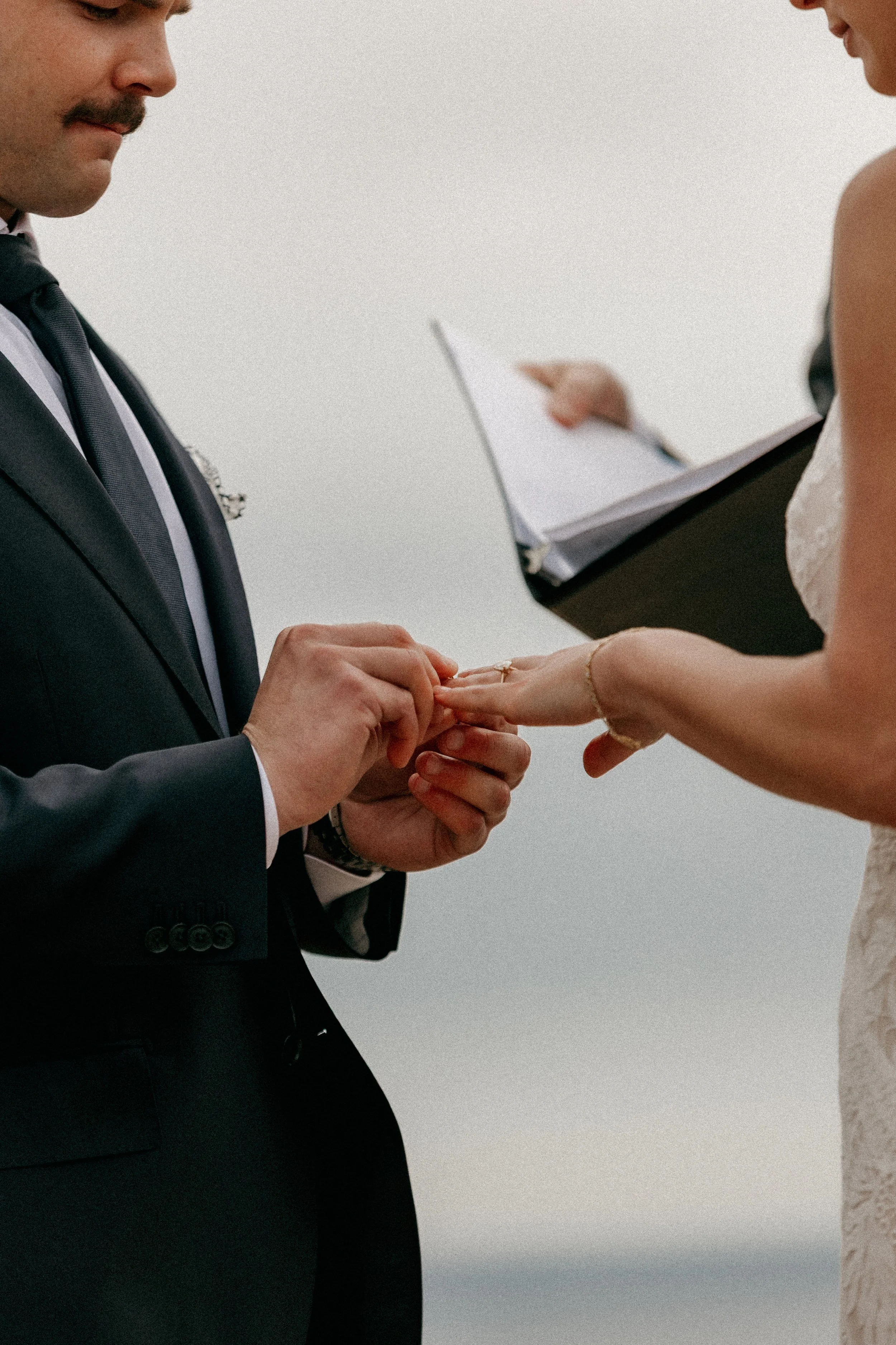 A groom in a black suit and tie is placing a ring on a bride's finger during a wedding ceremony. The woman is wearing a white lace wedding dress, and there is a person holding a clipboard and papers in the background.