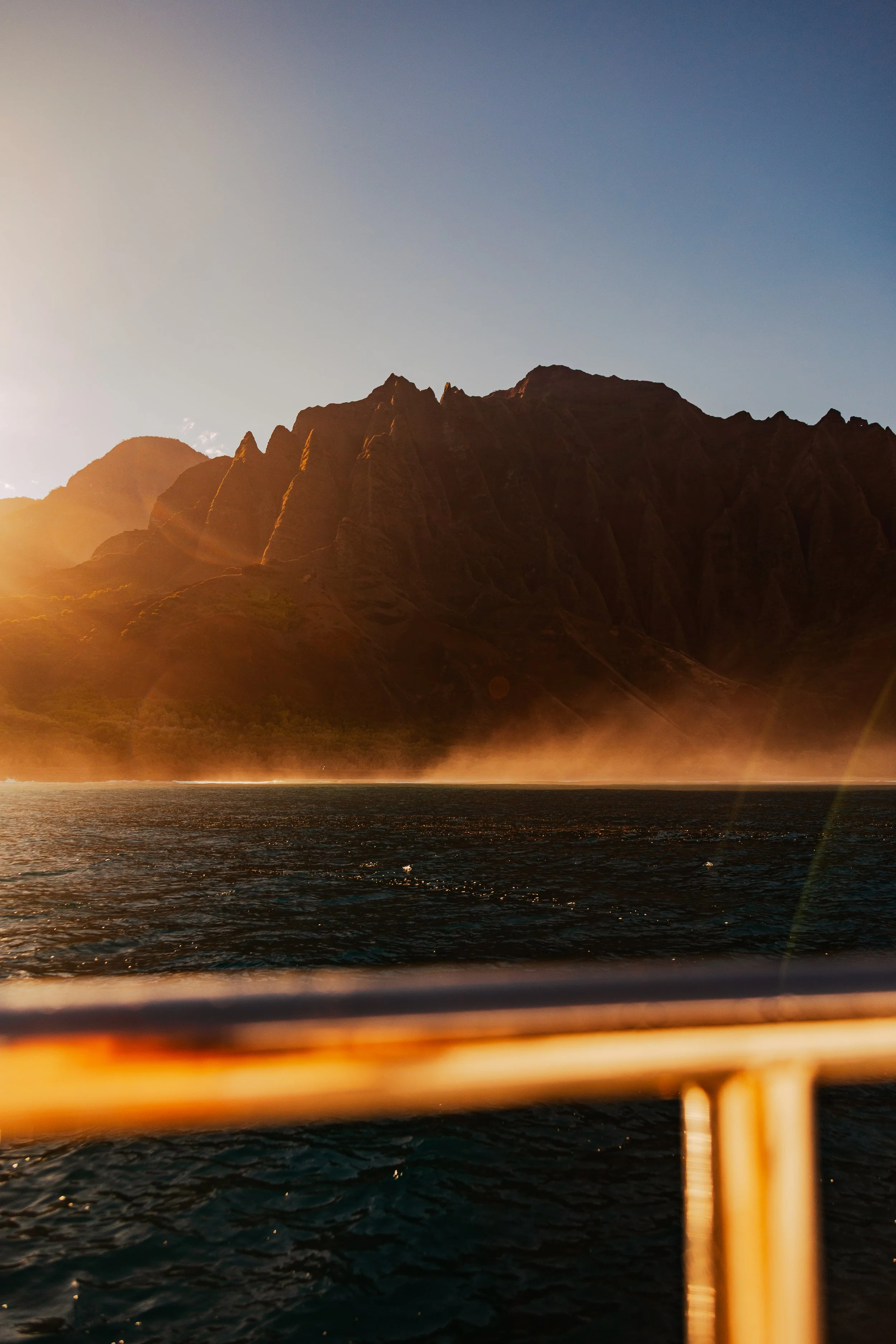 Sunset over mountainous coastline viewed from a boat, with a metal railing in the foreground.