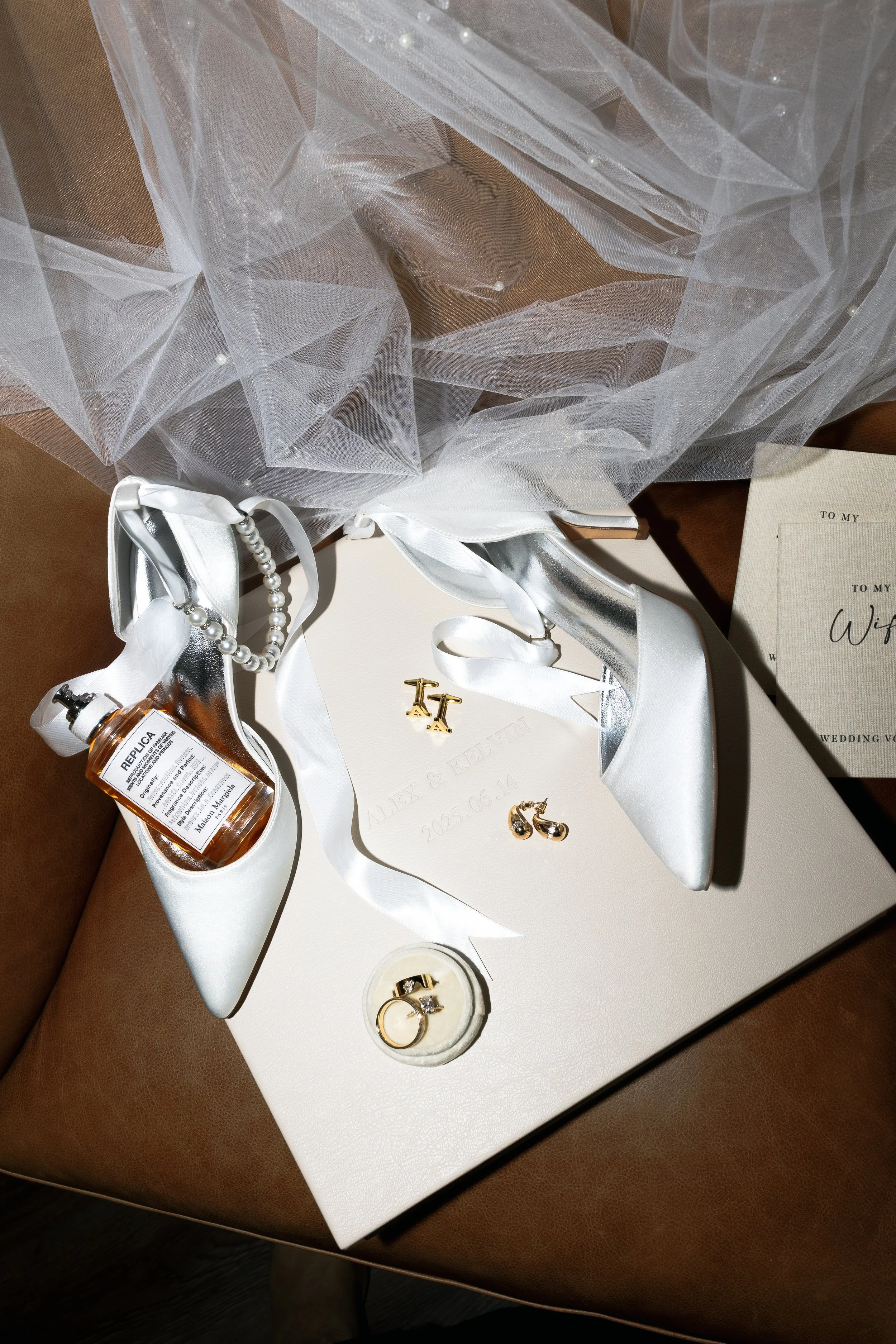 Wedding accessories including white satin shoes, a pearl necklace, gold earrings, a wedding ring, a ring box, a bottle of perfume, and engraved invitations, all arranged on a white card with wedding details, atop a brown sofa.