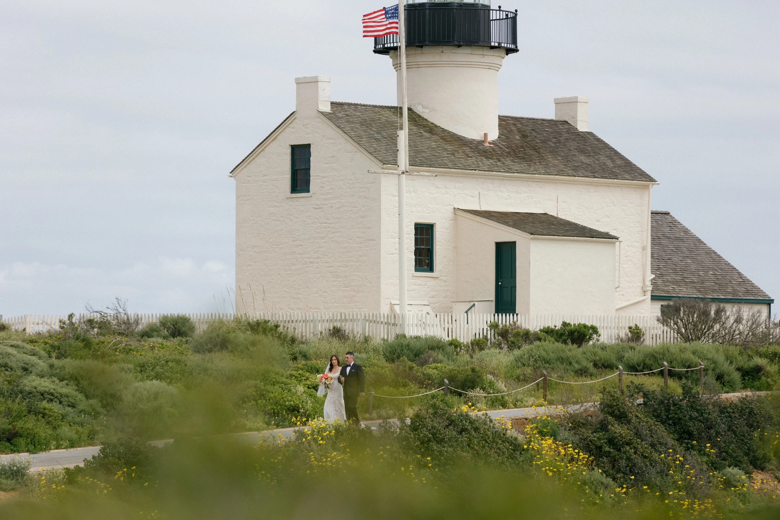 A bride and groom walking in front of a white lighthouse with a black top and an American flag, surrounded by greenery and yellow flowers.